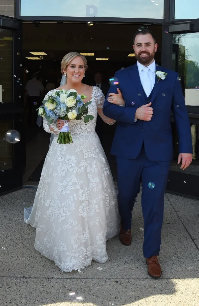 Bride Wearing Long Sleeve Floral Lace Wedding Dress Called Sawyer By Sottero and Midgley Holding Arm Of Groom With Bubbles In The Air