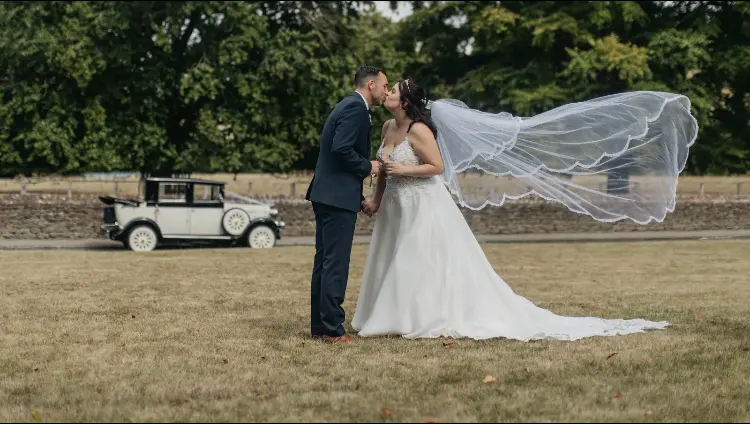 Bride Wearing Romantic Floral Wedding Dress With Lace Cutout Train Called Savannah By Maggie Sottero With Groom And Flying Veil And Vintage Car