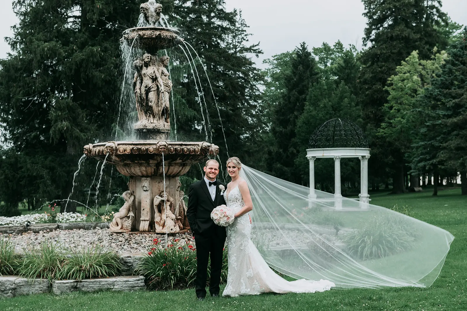 Newlyweds stood by large Roman style water fountain smiling with brides veil floating in the breeze.