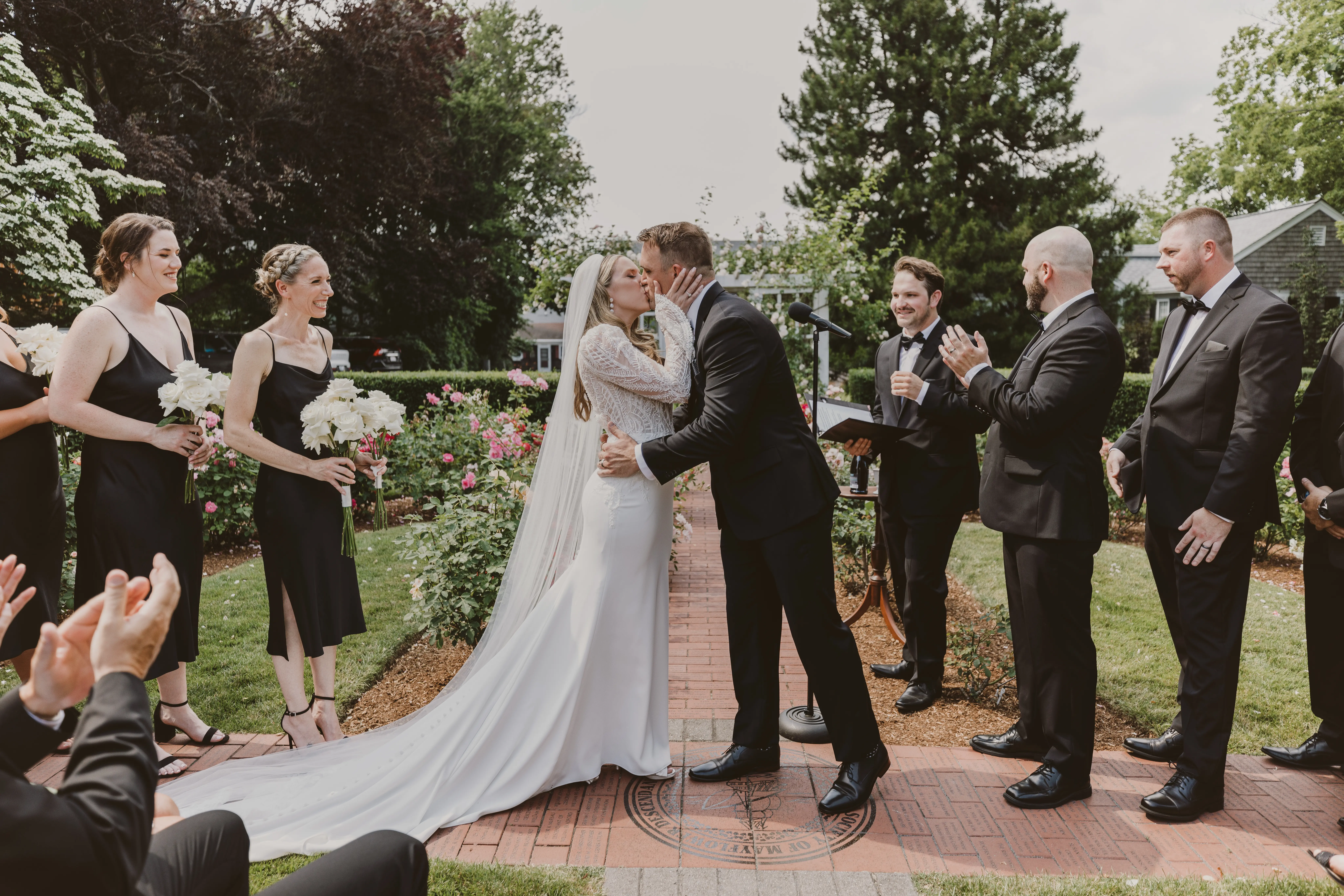 Bride and groom kissing while wedding party, all dressed in black, clap and cheer.