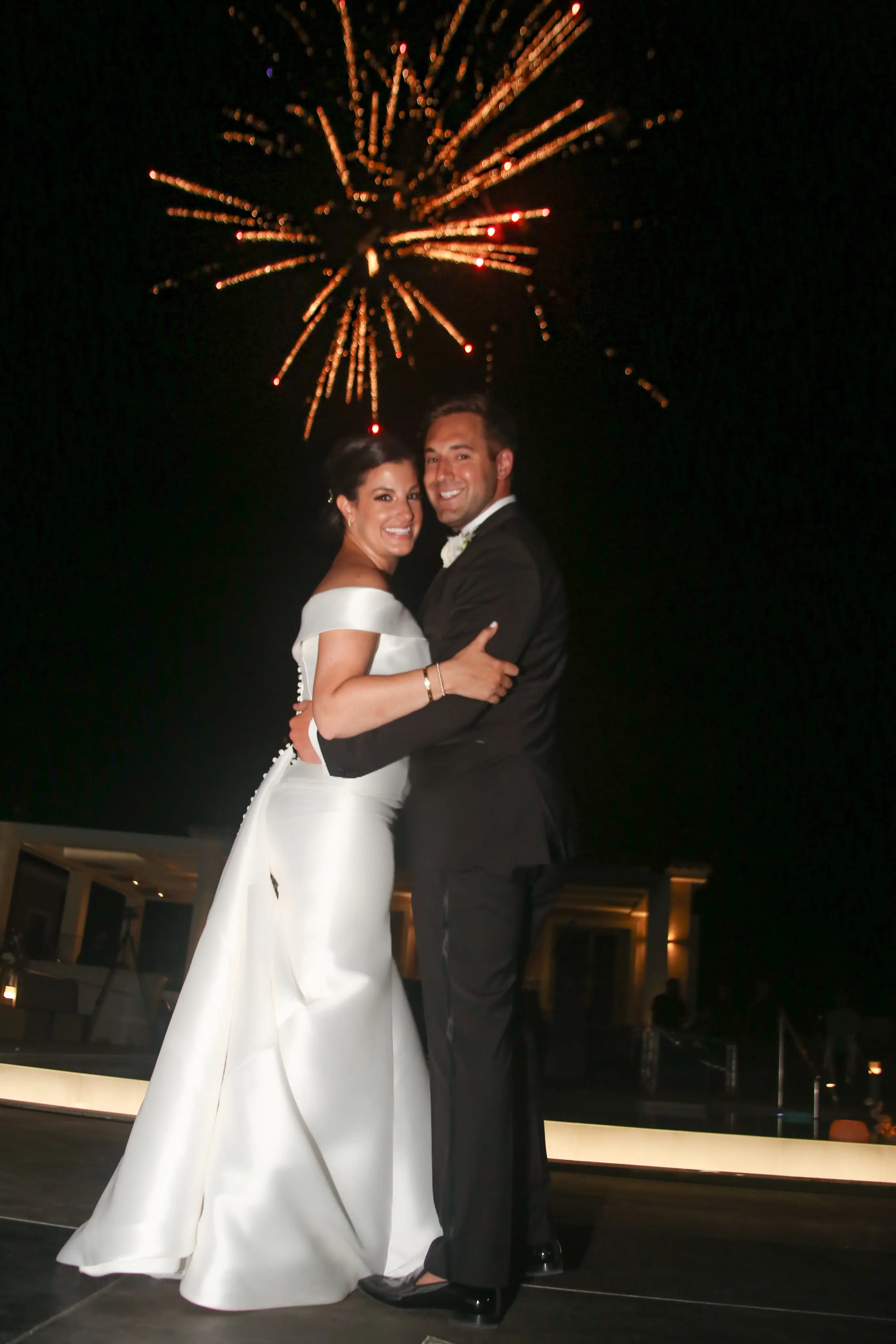 Bride wearing Simple Fit-and-Flare Wedding Dress called Josie by Rebecca Ingram kissing the groom with fireworks behind them