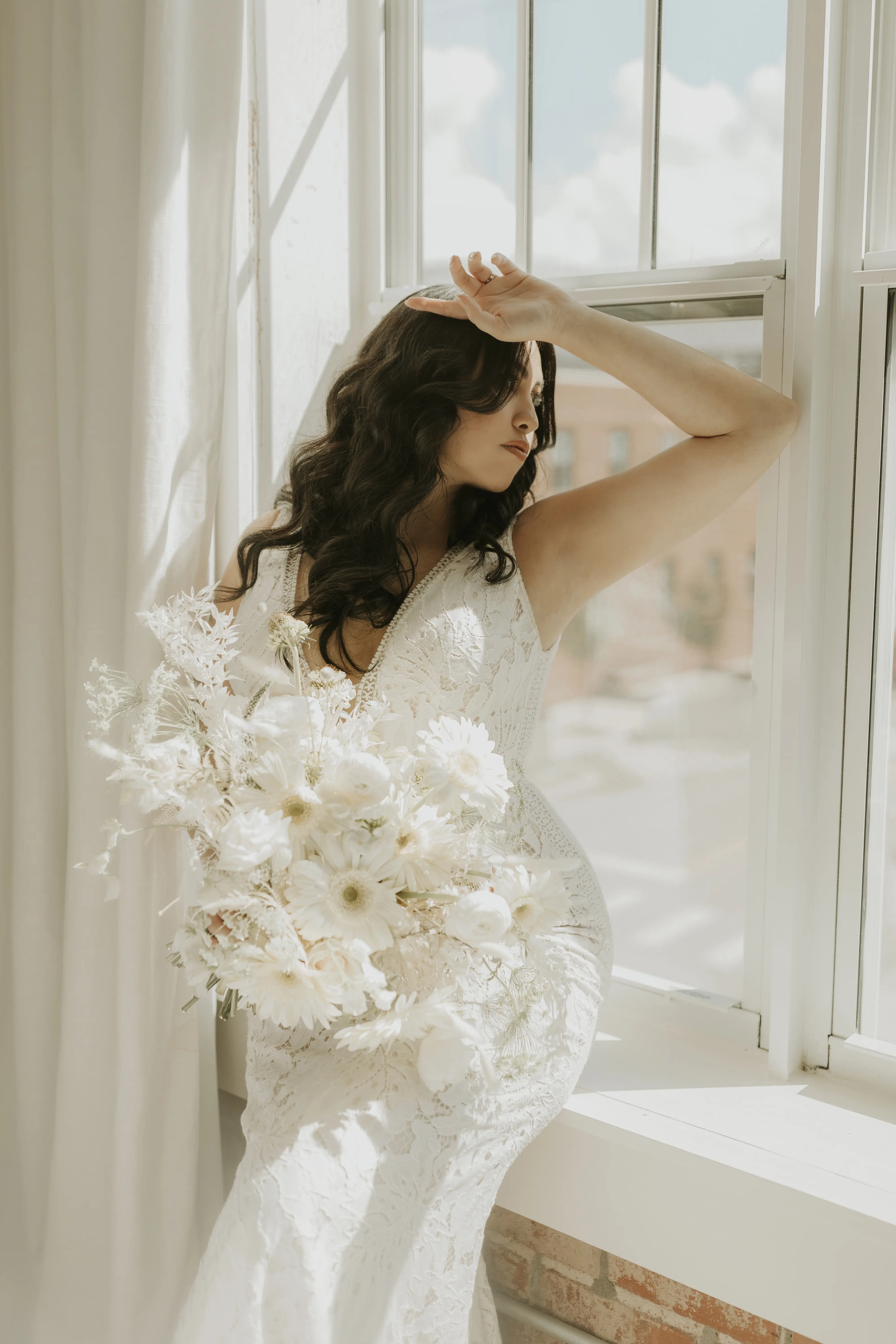 Bride wearing wedding dress called Burke holding all white/ivory bouquet gazing out sunny window.