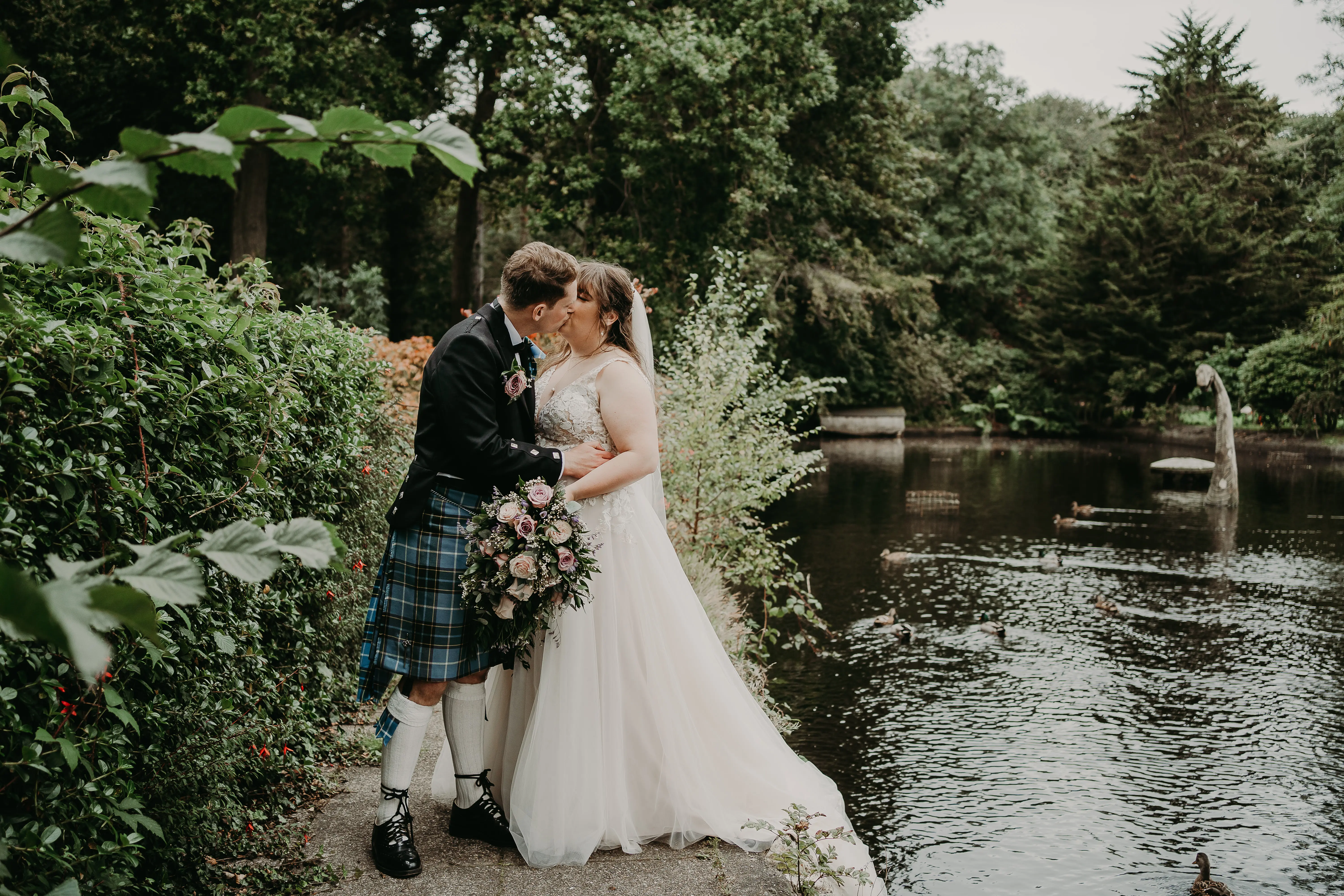 Bride and groom kissing near lake in Scotland with faux loch ness monster in background.