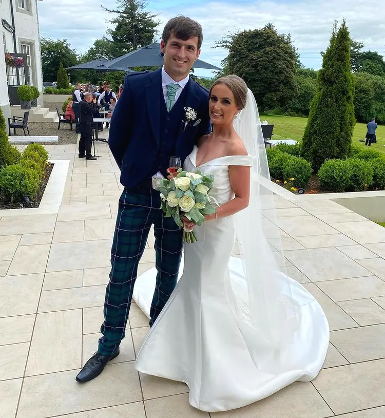 Groom stood with bride wearing wedding dress called Josie and holding ivory roses.