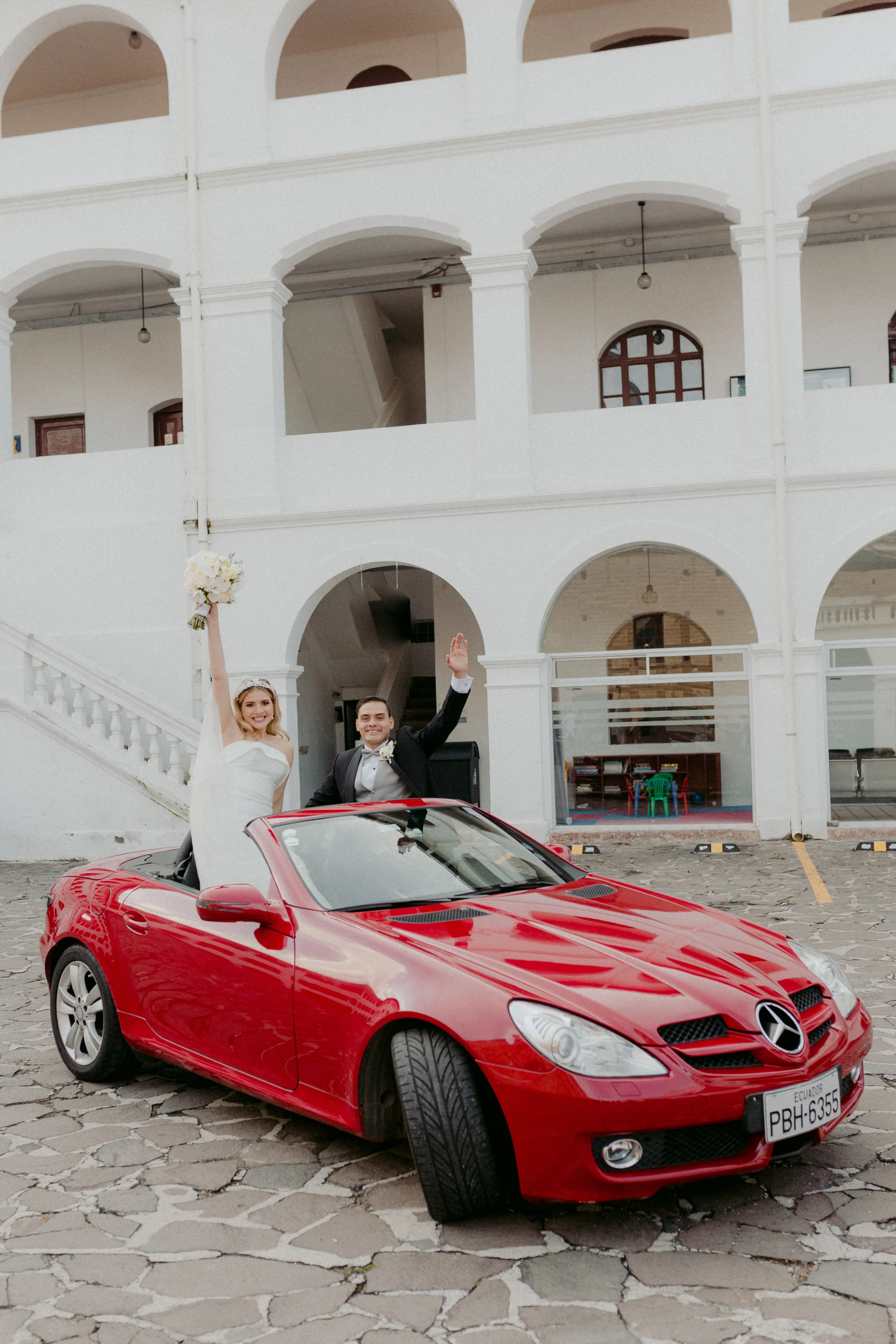 Bride Holding Up Flower Bouquet in a Red Car with Groom Wearing Wedding Dress Called Mitchell by Maggie Sottero