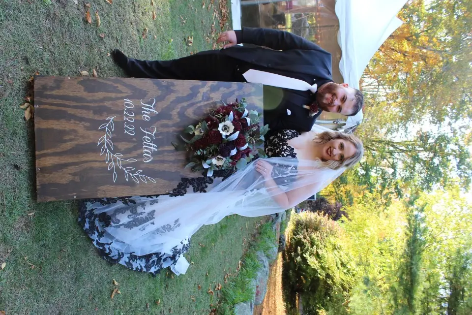 Bride Wearing Black Lace Wedding Dress Called Tristyn By Maggie Sottero With Groom Next To Their Wedding Sign