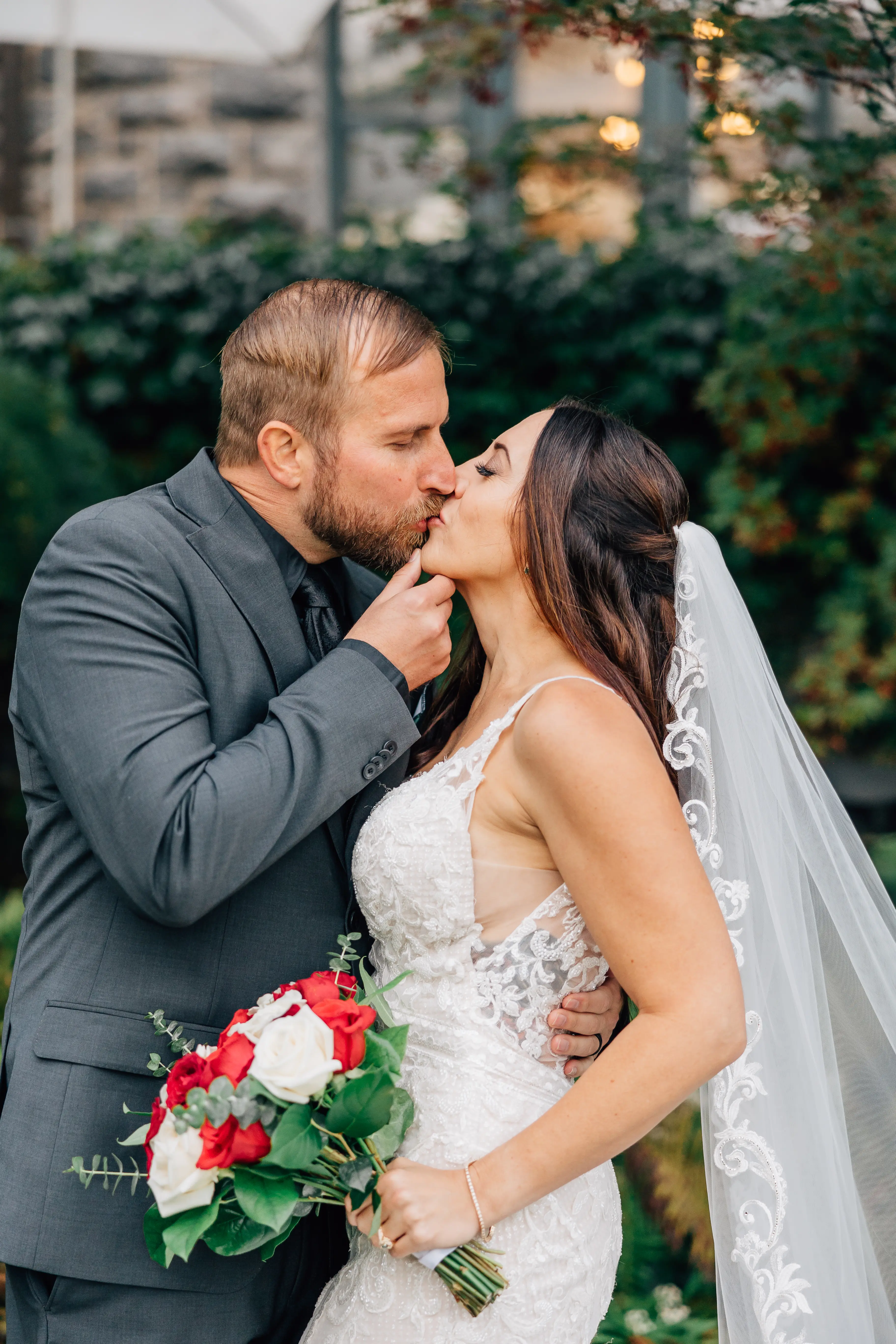 Groom Kissing Bride Wearing A Fitted Lace Wedding Dress Named Veronique by Maggie Sottero With A Veil And Bouquet Of Red And White Roses