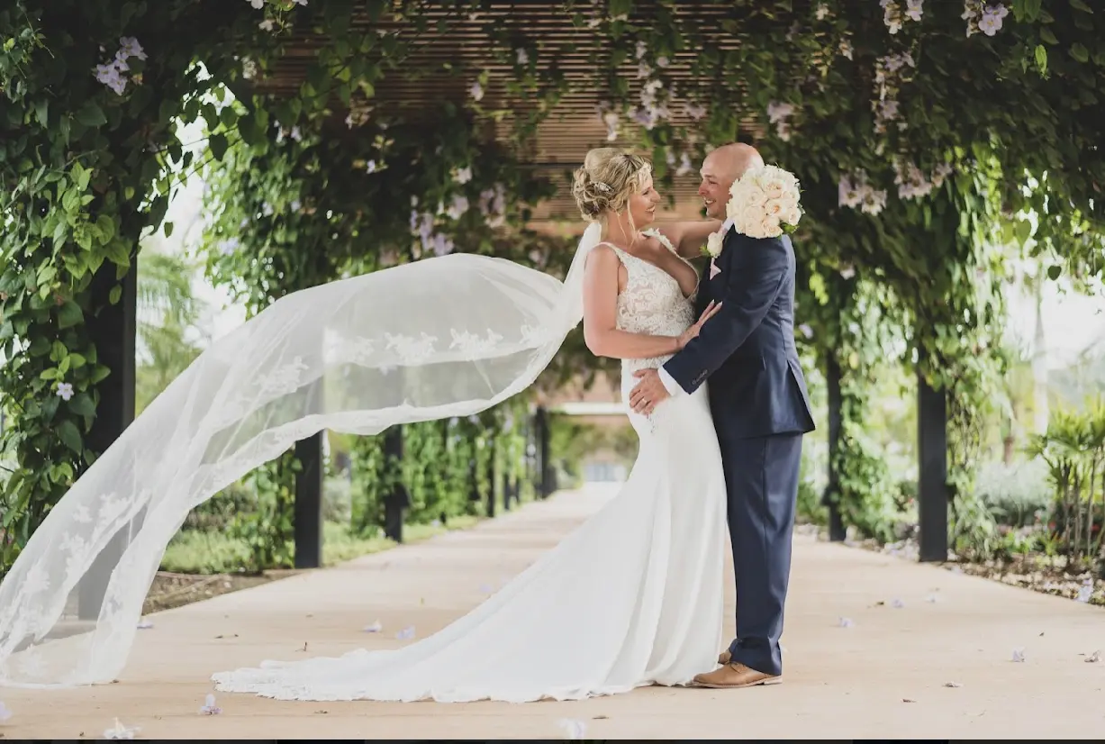 Groom and bride posing under tunnel of trees and blooms.