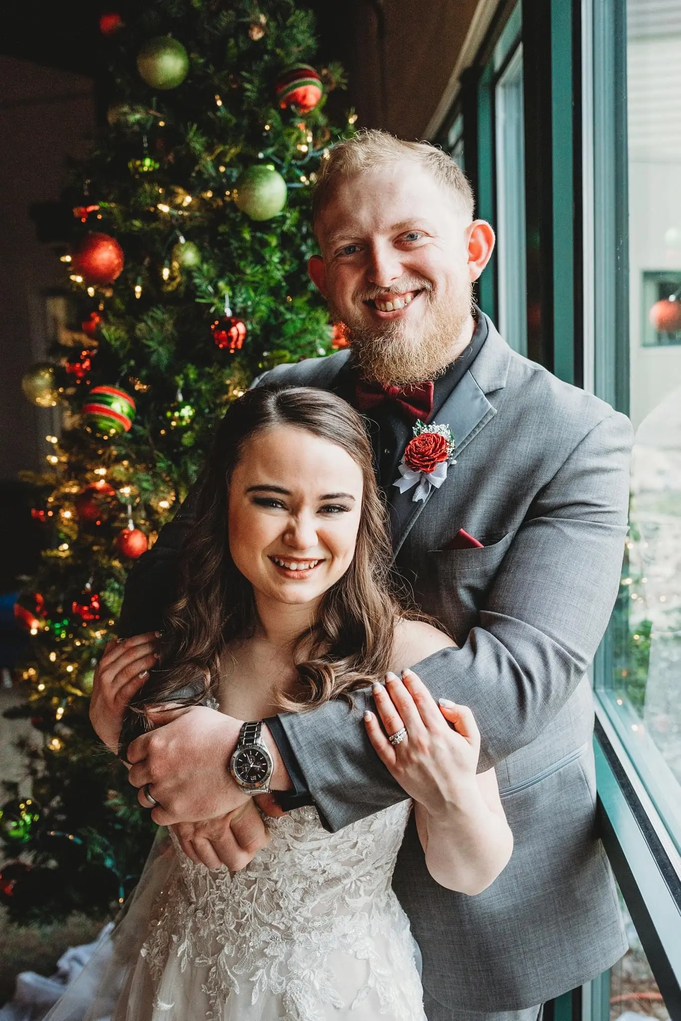 Bride And Groom Standing In Front Of Christmas Tree. Bride Wearing Lace Sparkly Wedding Dress Called Stevie By Maggie Sottero