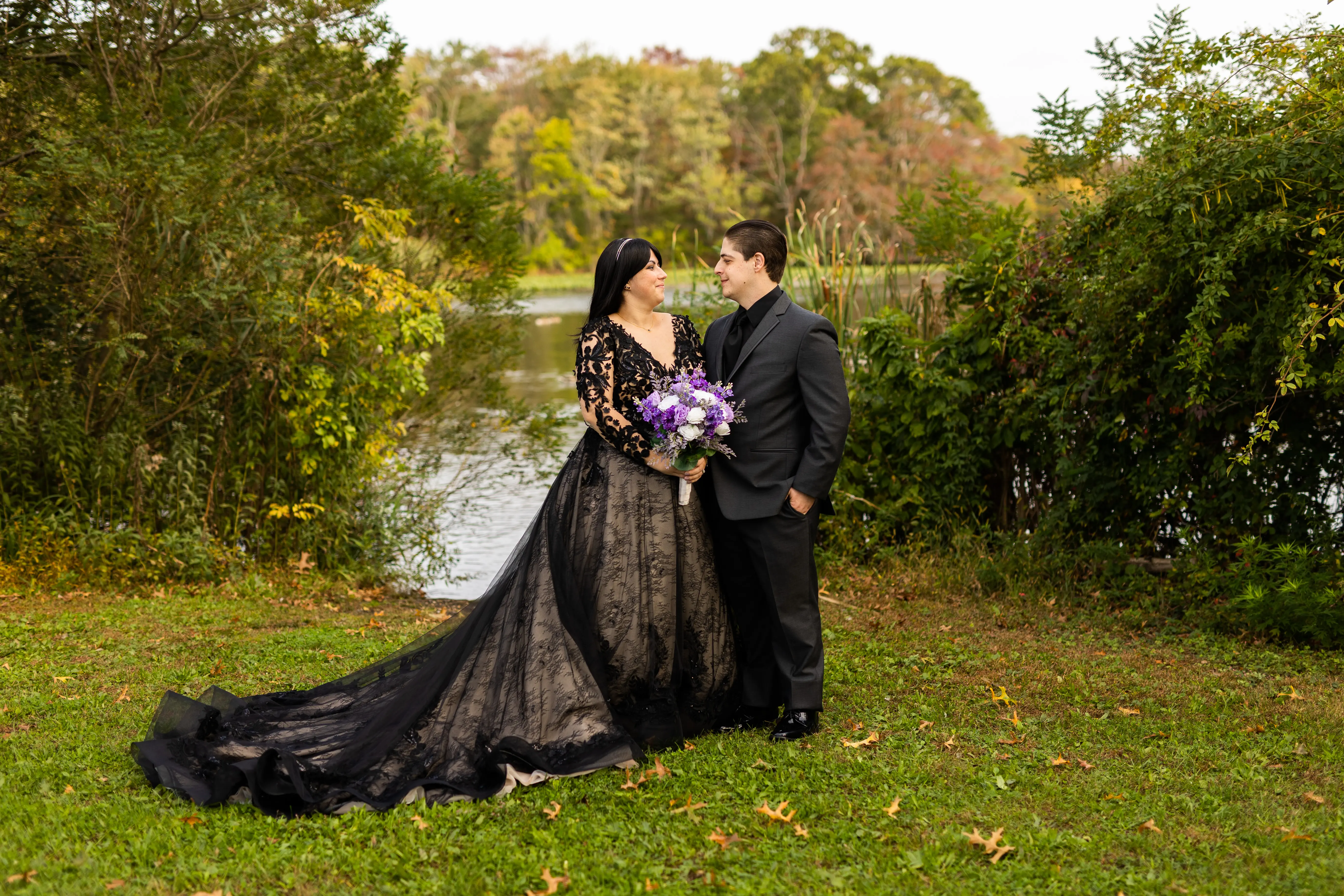 Bride in a Field with Groom Wearing Long Sleeve Black Lace Ballgown Wedding Dress Holding Bridal Bouquet.