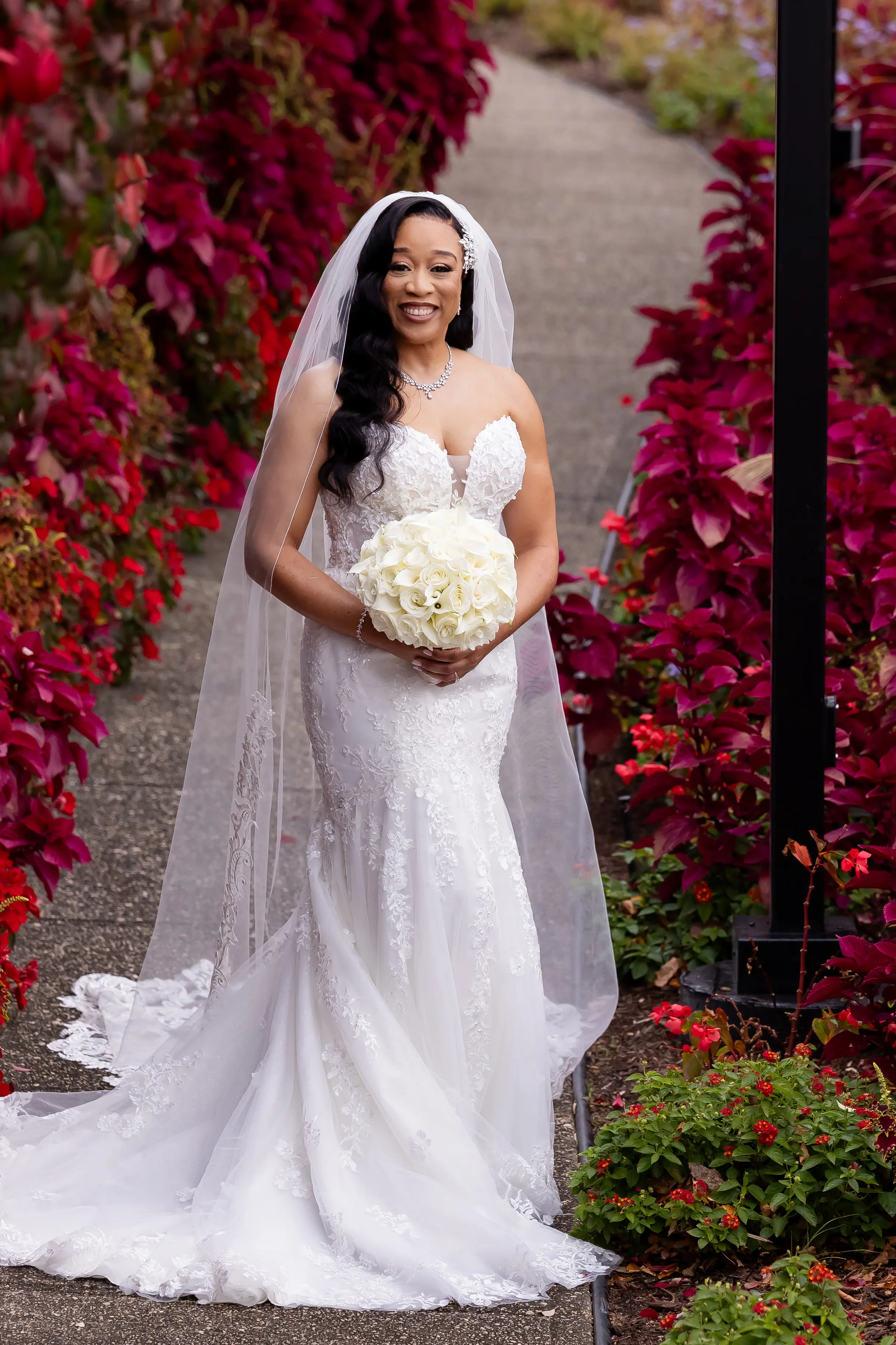 Bride Wearing Sexy Lace Wedding Dress Called Lennon By Maggie Sottero Next To Red Floral Bushes