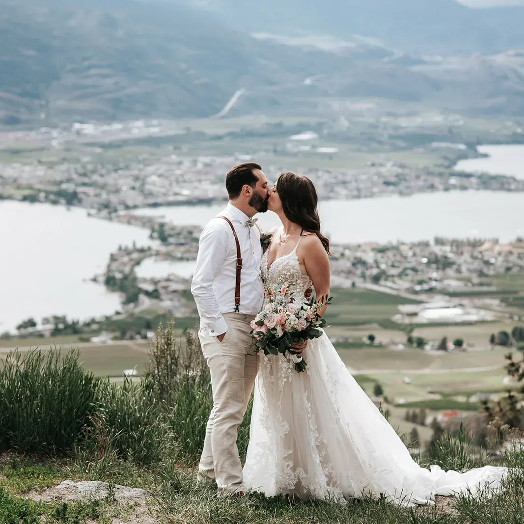 Bride Wearing Romantic Key Hole Back A-Line Wedding Dress Called Marlow By Maggie Sottero Kissing Groom Overlooking A Town