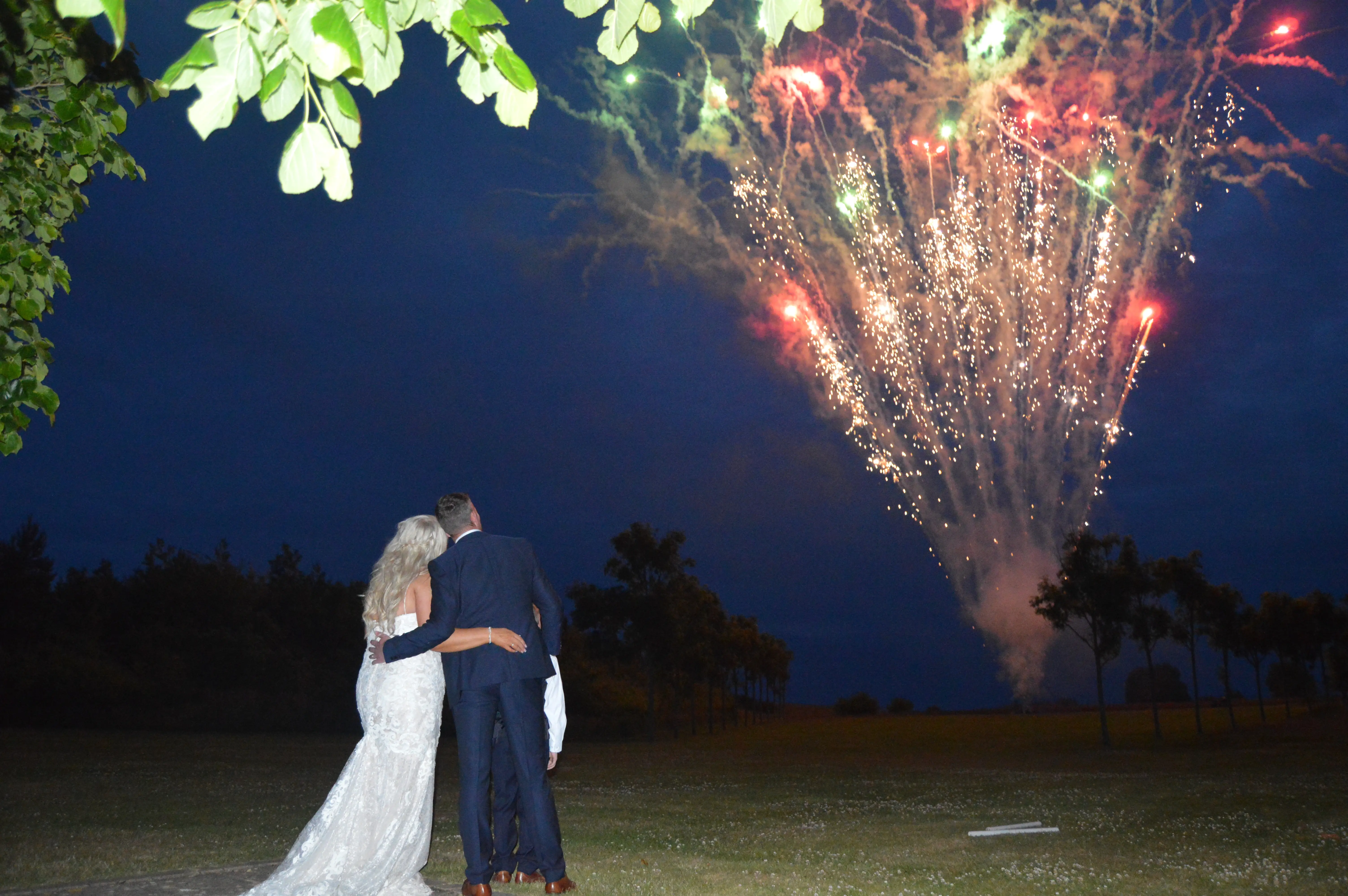 Newlyweds embrace each other as they watch red and gold fireworks in the sky.