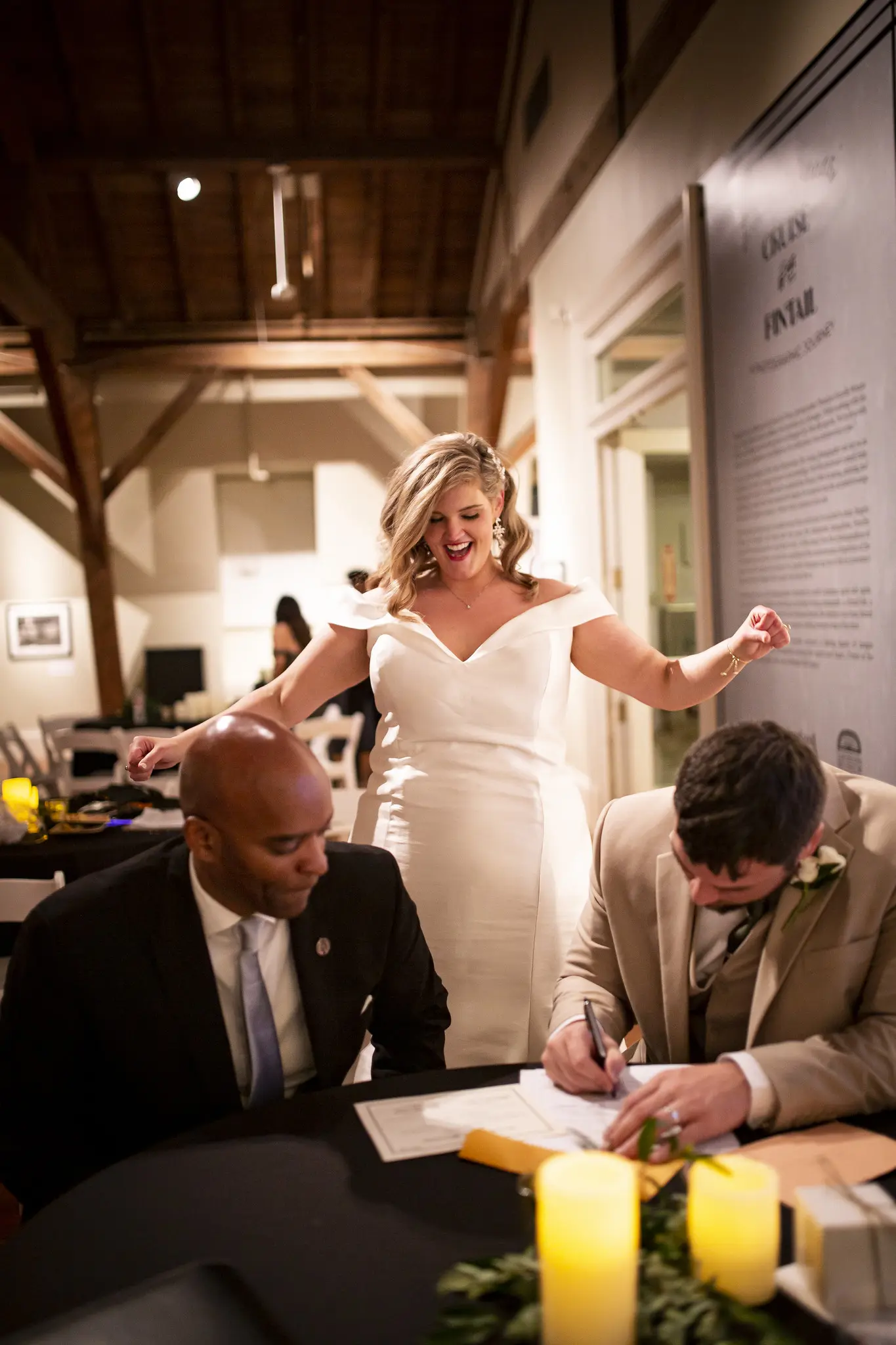 Couple Signing Marriage Papers. Bride Wearing Vintage Glamour Timeless Off-The-Shoulder Fit-And-Flare Wedding Dress Called Josie By Rebecca Ingram