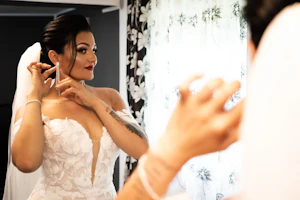 Bride Wearing Lace Mermaid Wedding Gown Called Hattie By Rebecca Ingram Admiring Her Earrings In A Mirror