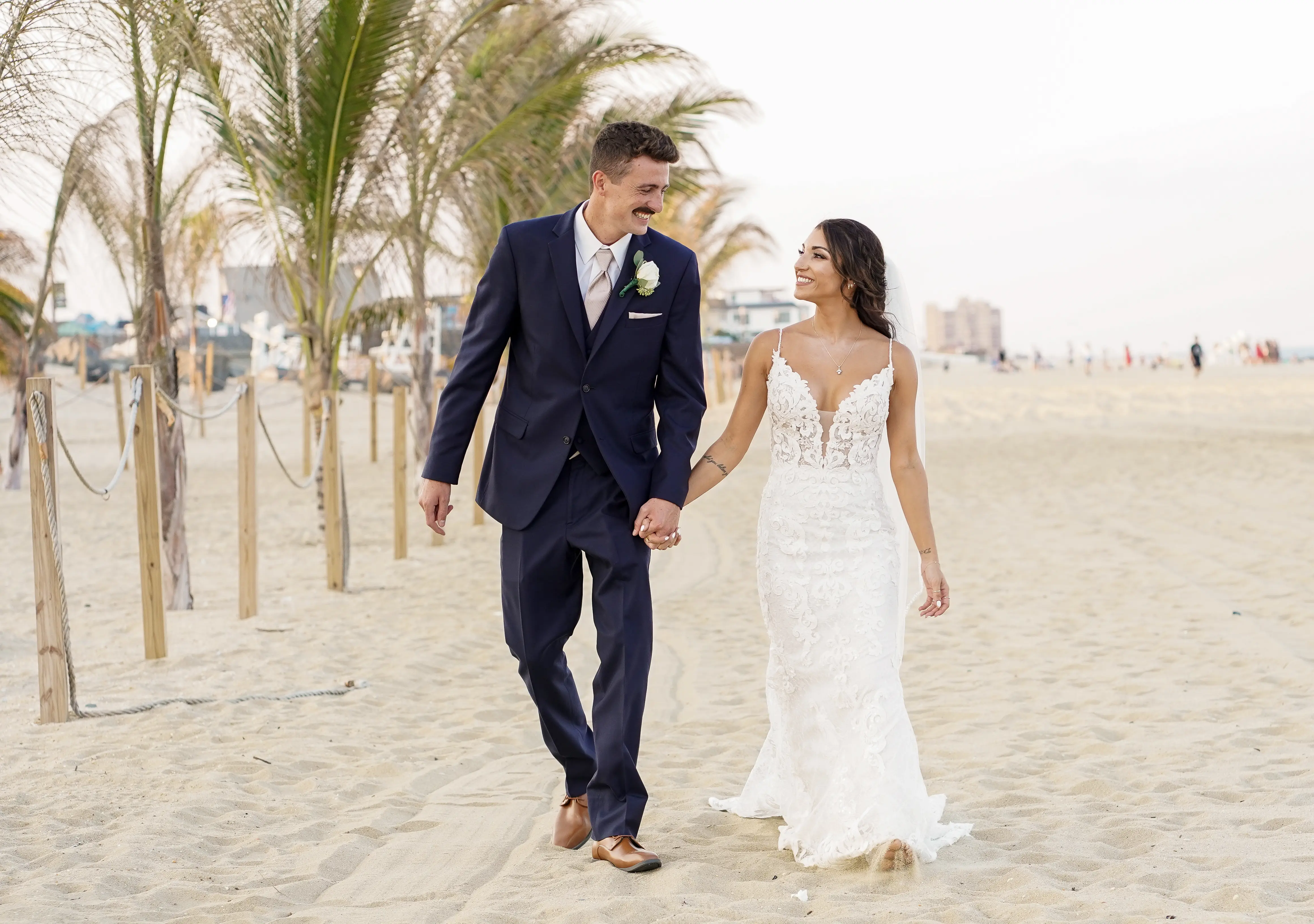 Groom Holding Hands with Bride Walking on Beach Wearing Wedding Dress called Tuscany by Maggie Sottero.