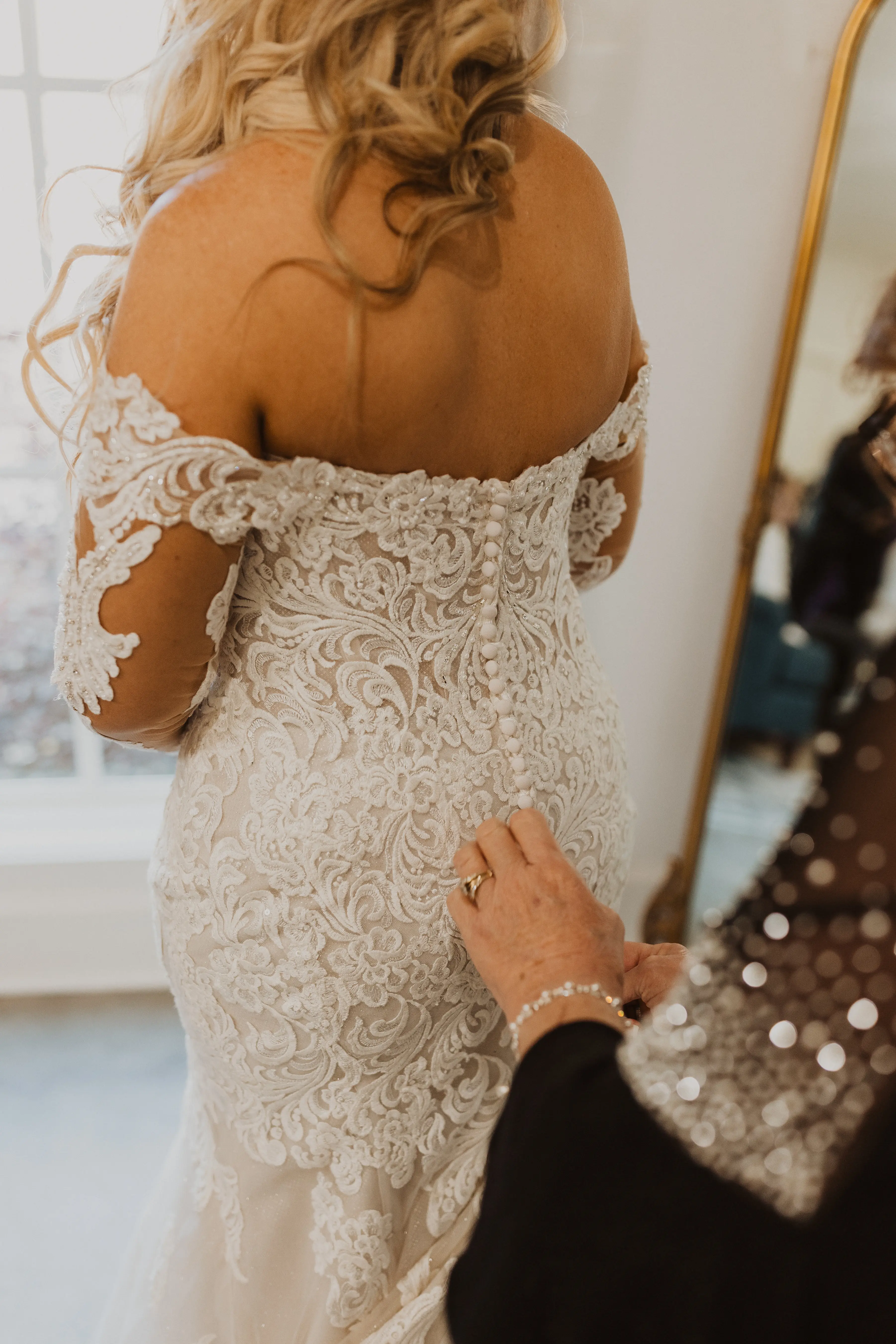 Bride In A Sparkly Lace White Wedding Dress Named Fiona By Maggie Sottero With Someone Buttoning Up The Back Of The Dress