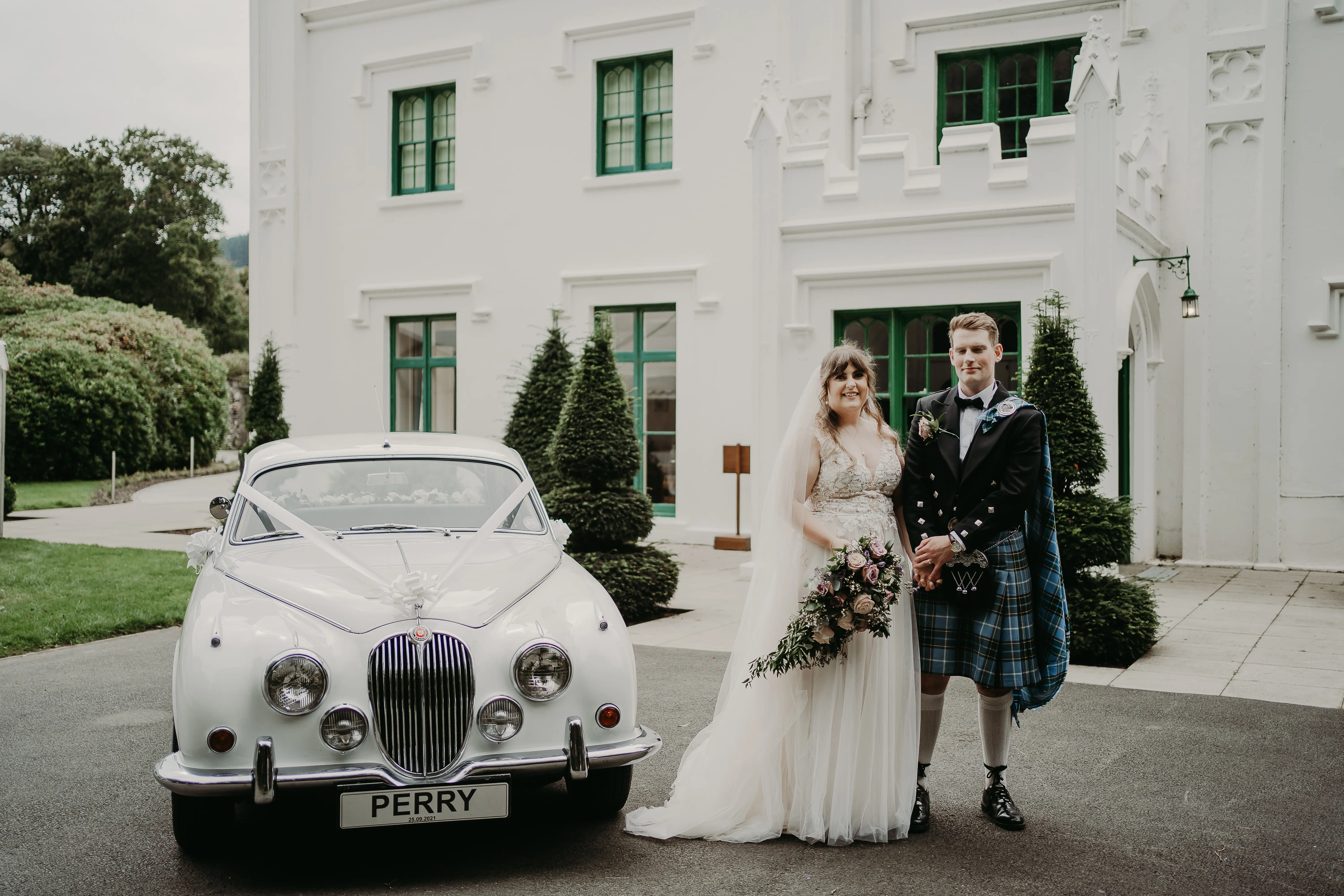 Bride wearing wedding dress called Minerva and groom wearing kilt stood by vintage car.