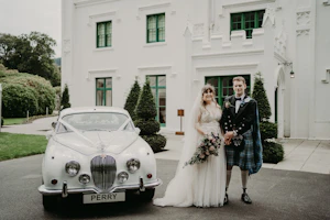 Bride wearing wedding dress called Minerva and groom wearing kilt stood by vintage car.