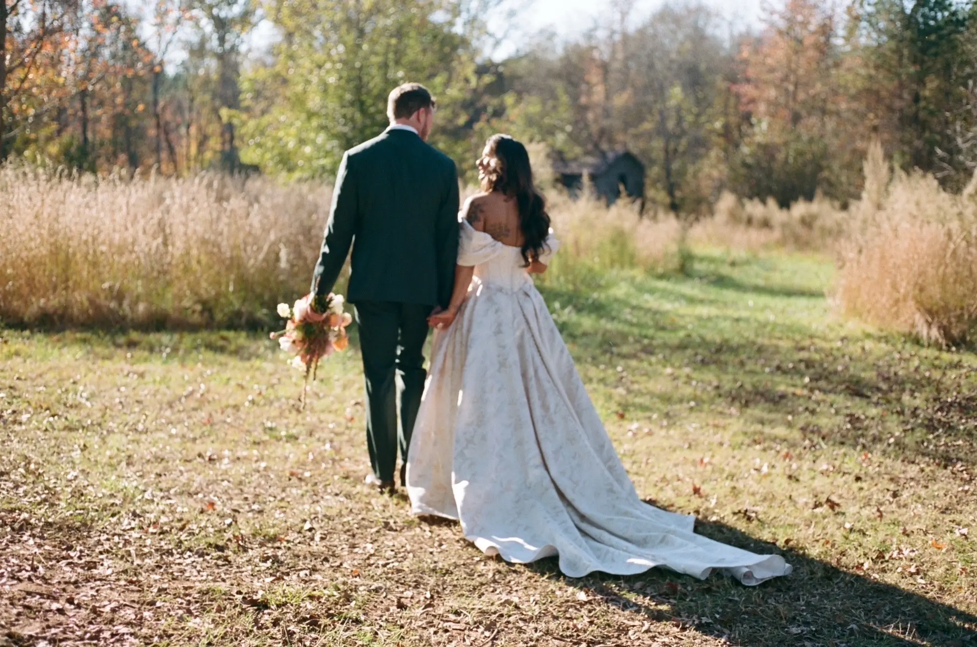 Bride Wearing Floral Ballgown Wedding Dress Called Derrick Marie Holding Hands Walking With Groom In A Field