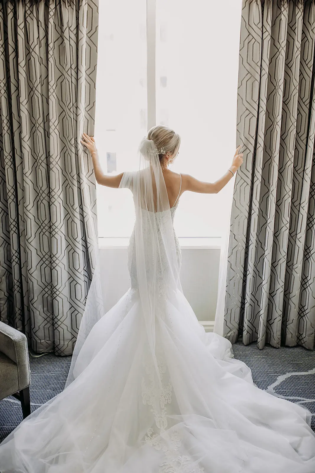 Back view of bride opening curtains wearing wedding dress called Alistaire.
