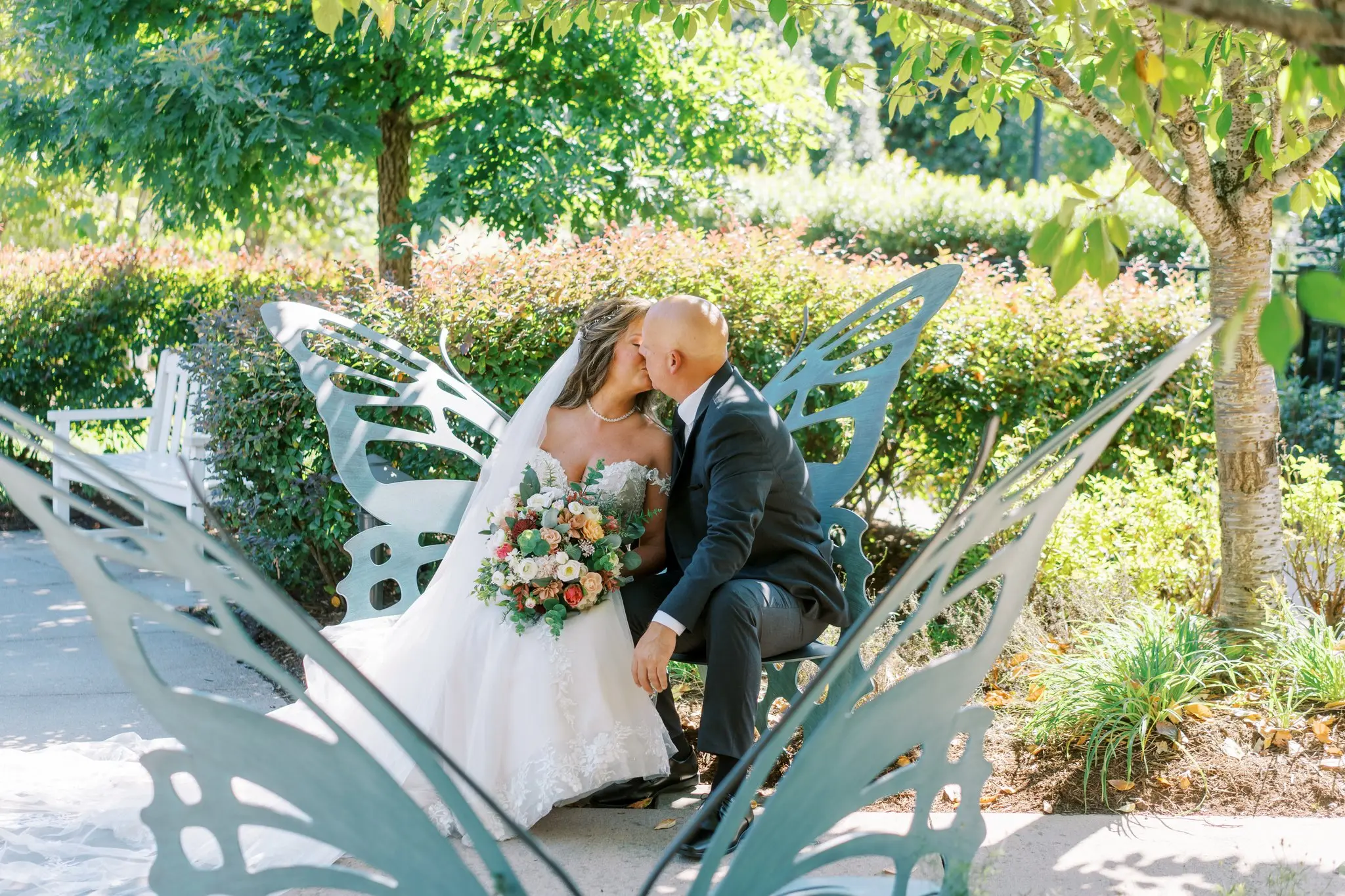 Bride Wearing Sexy Lace Wedding Dress Called Lennon By Maggie Sottero Kissing Groom On A Butterfly Bench