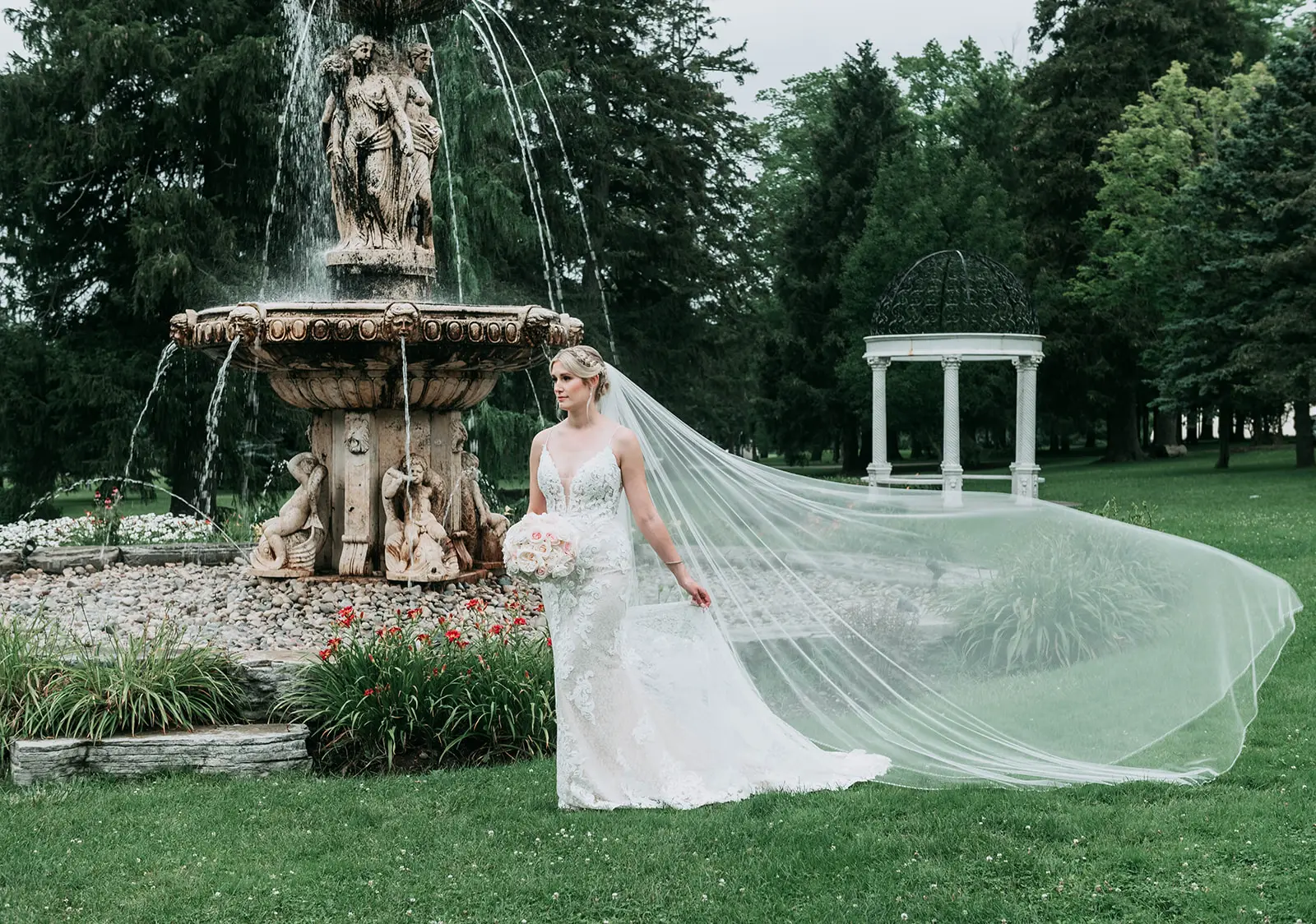 Bride stood by Roman style fountain with veil blowing in the wind.