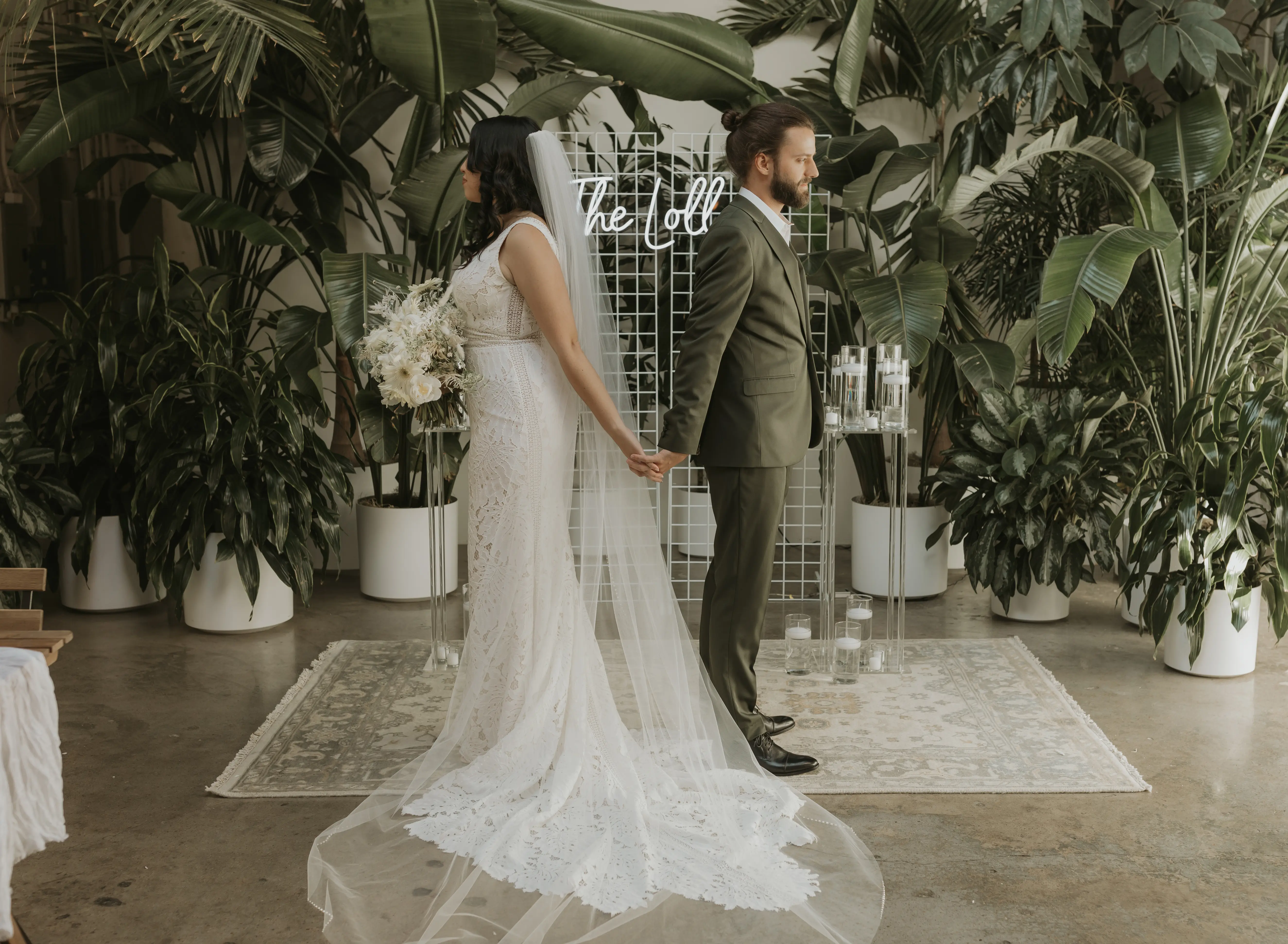 Bride and groom back facing holding hands with lots of greenery in background.