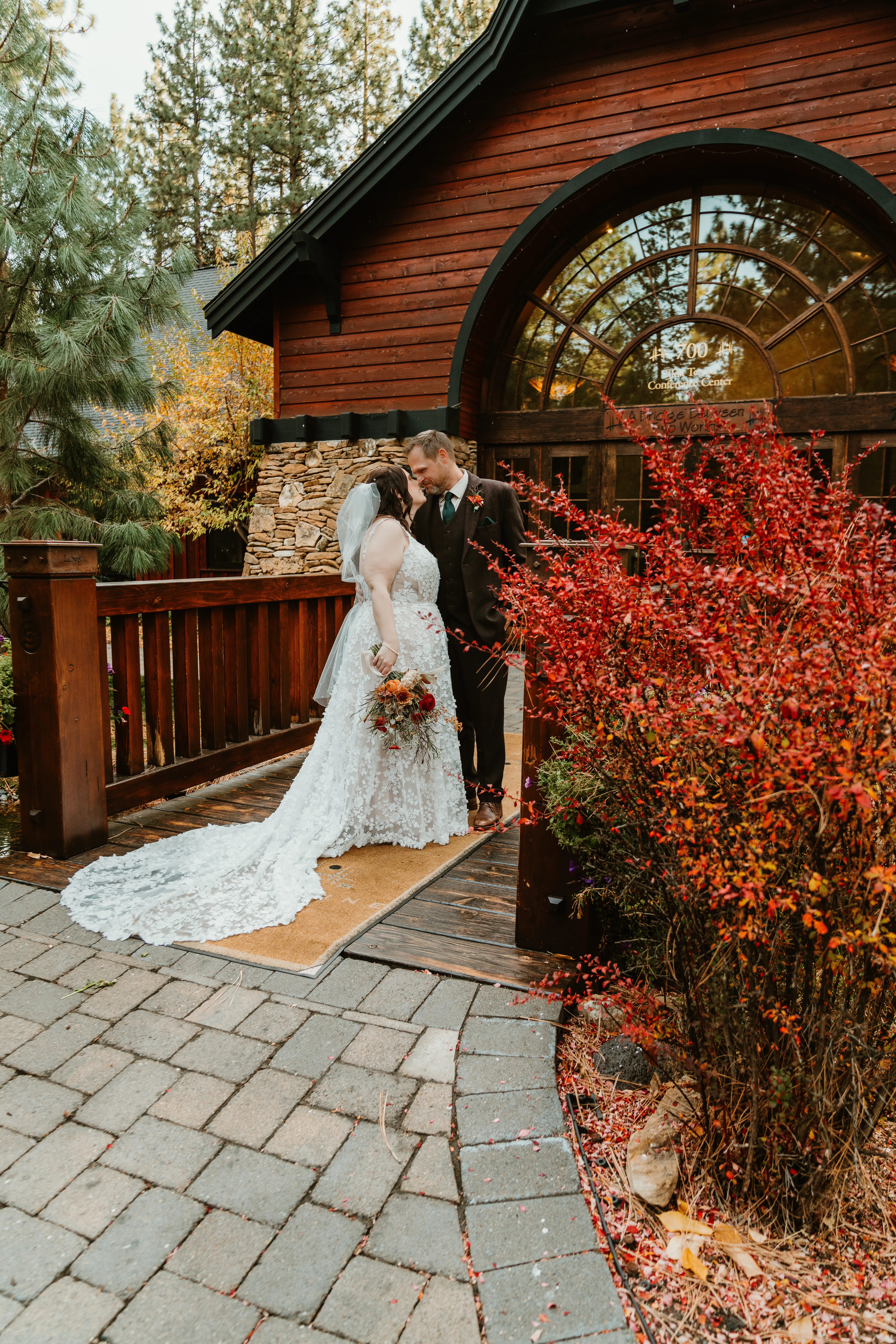 Bride Wearing Lace Wedding Dress Called Ladonna By Maggie Sottero Kissing Groom In Front Of A Cabin
