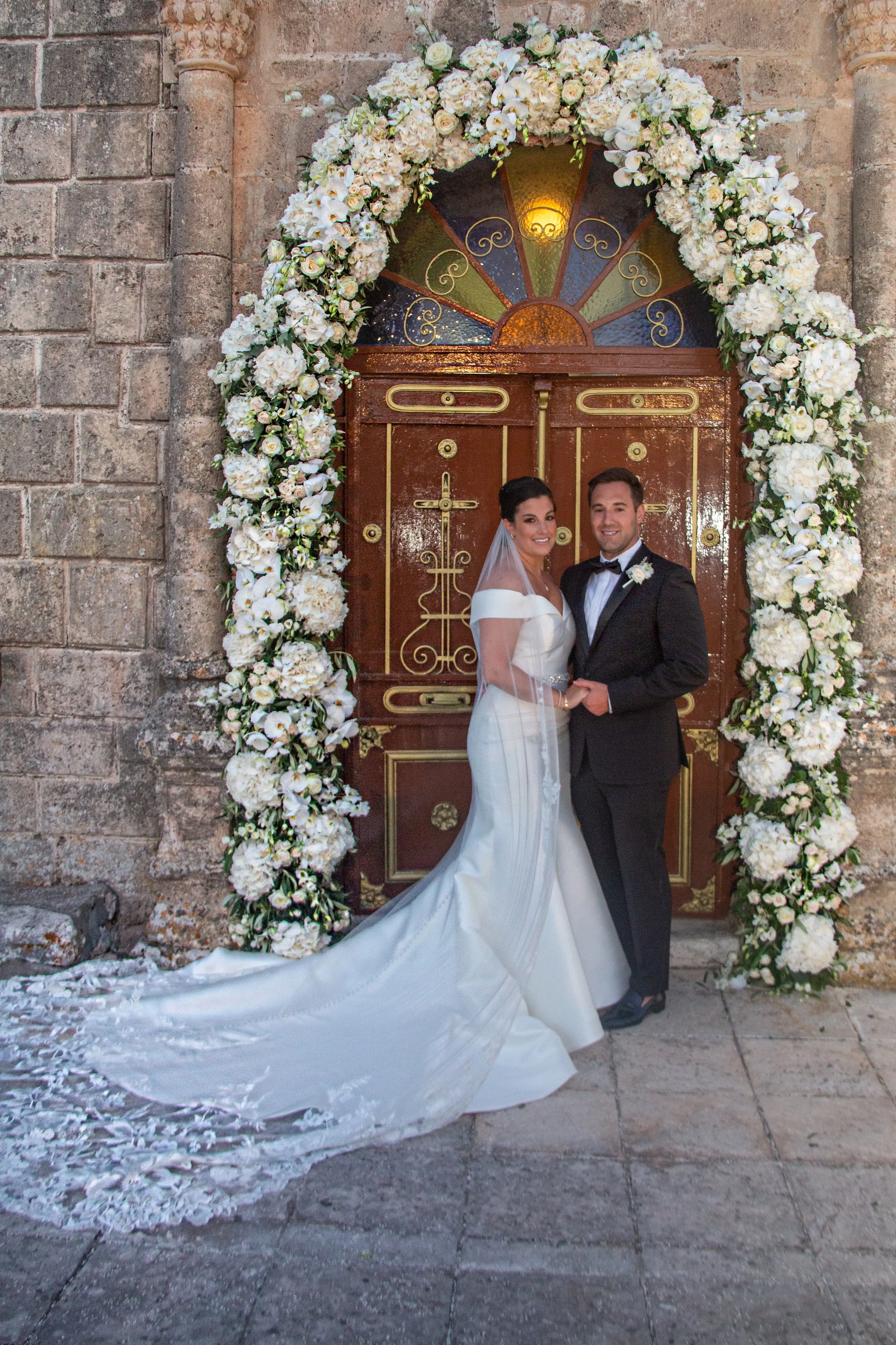 Bride wearing Simple Fit-and-Flare Wedding Dress called Josie by Rebecca Ingram under a flower arch with the groom