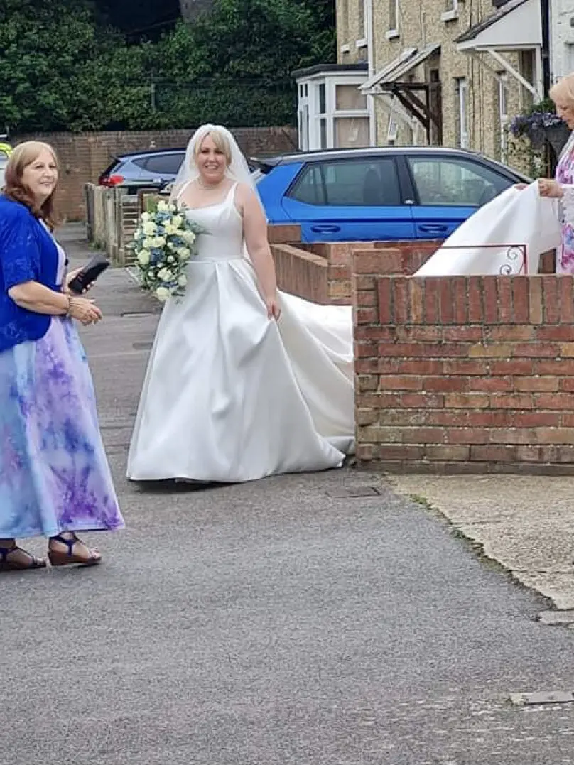Bride leaving her house on wedding day with family member holding her train.