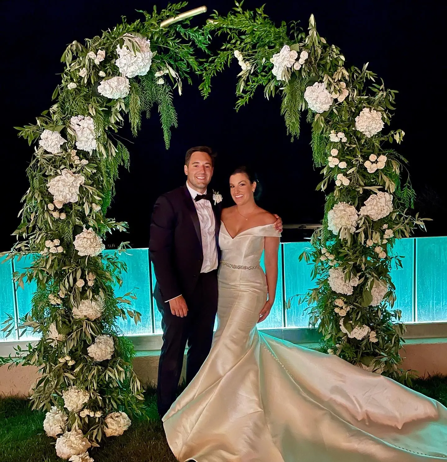 Bride wearing Simple Fit-and-Flare Wedding Dress called Josie by Rebecca Ingram under a flower arch with the groom