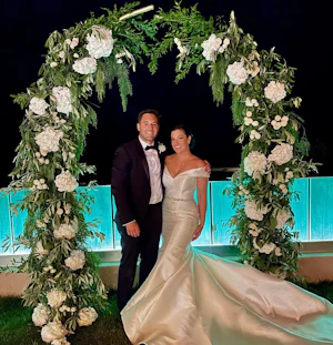 Bride wearing Simple Fit-and-Flare Wedding Dress called Josie by Rebecca Ingram under a flower arch with the groom
