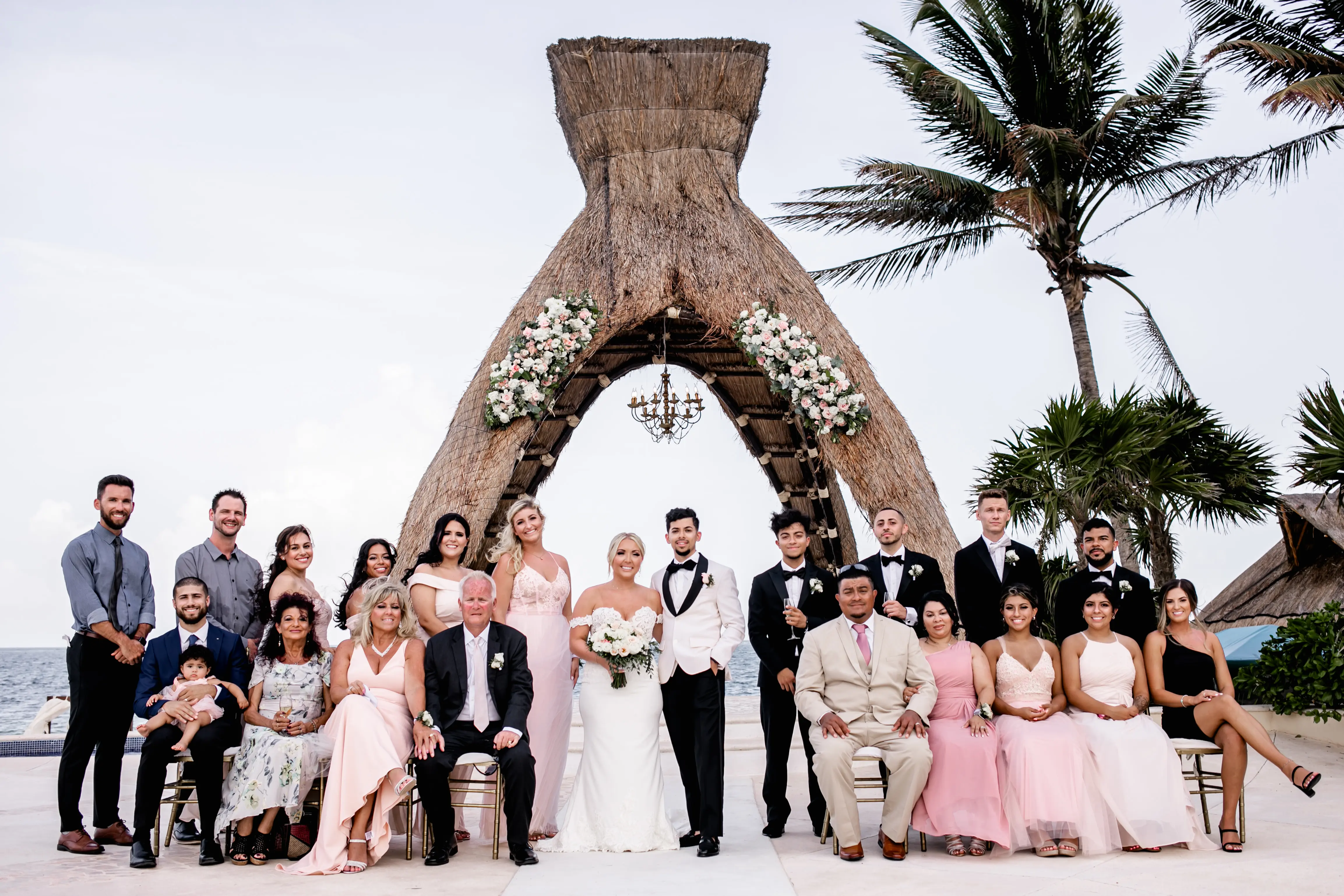 Wedding party with bride and groom stood under straw archway.