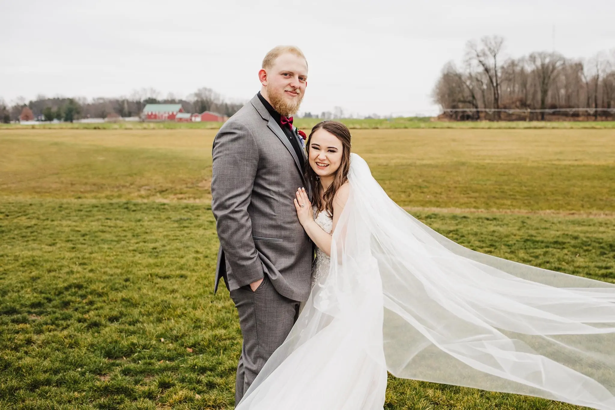 Bride And Groom Standing Together In A Field. Bride Wearing Lace Tulle Wedding Dress Called Stevie By Maggie Sottero