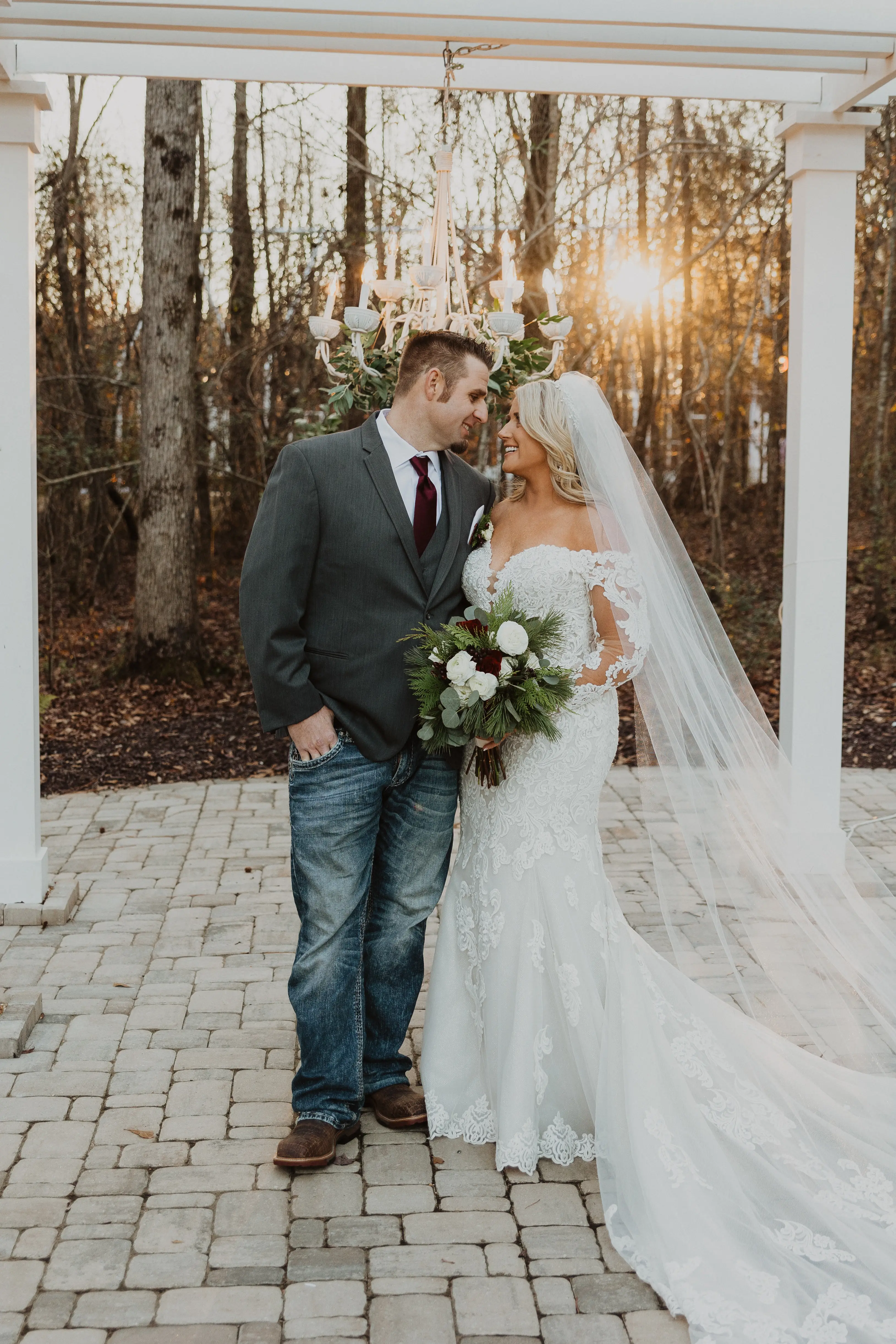 Groom Smiling With Bride In A White Wedding Dress With Off-The-Shoulder Sleeves Named Fiona By Maggie Sottero With A Long Veil And Flower Bouquet