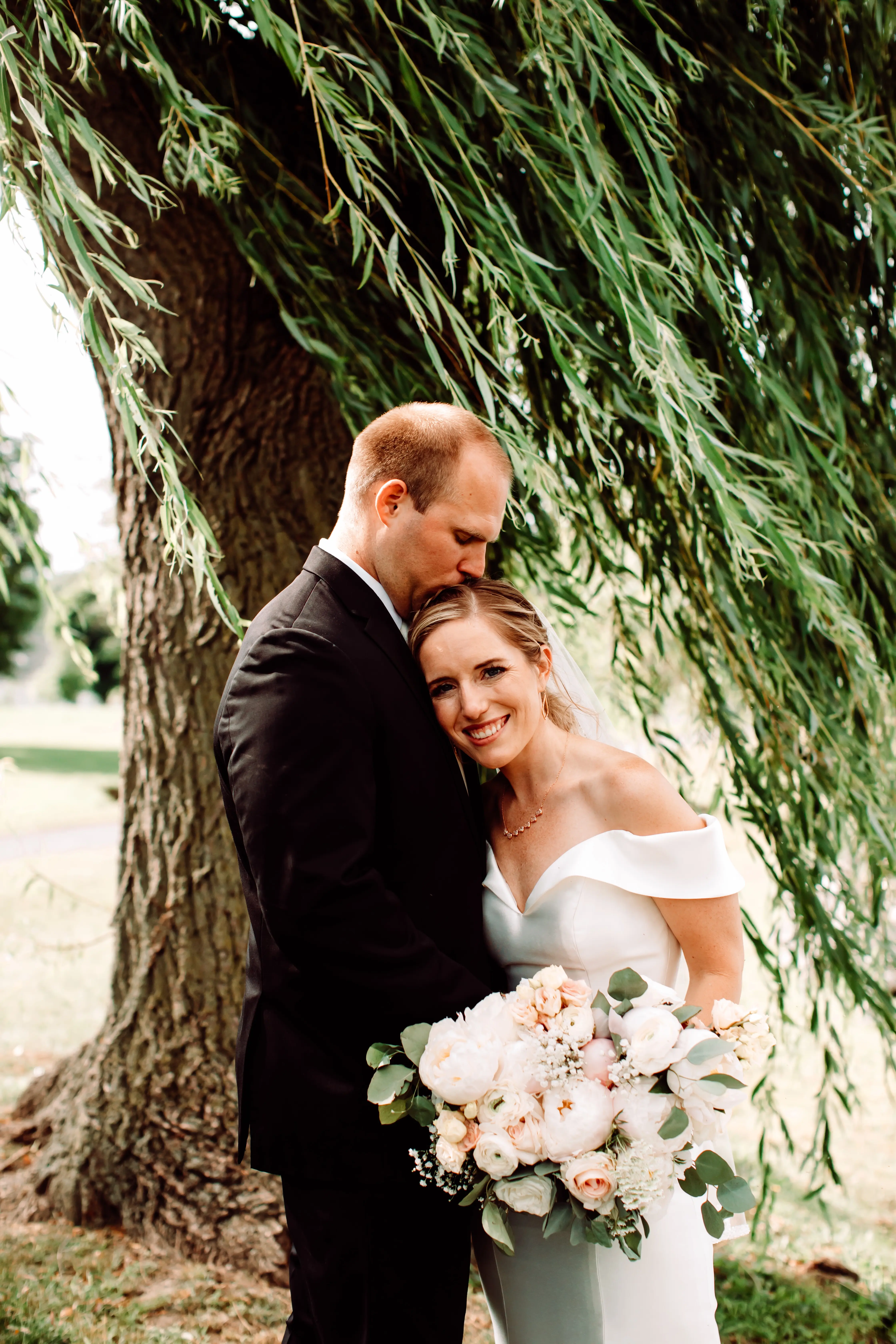 Groom kissing bride on the head as she smiles holding gorgeous peachy bouquet wearing wedding dress called Josie.