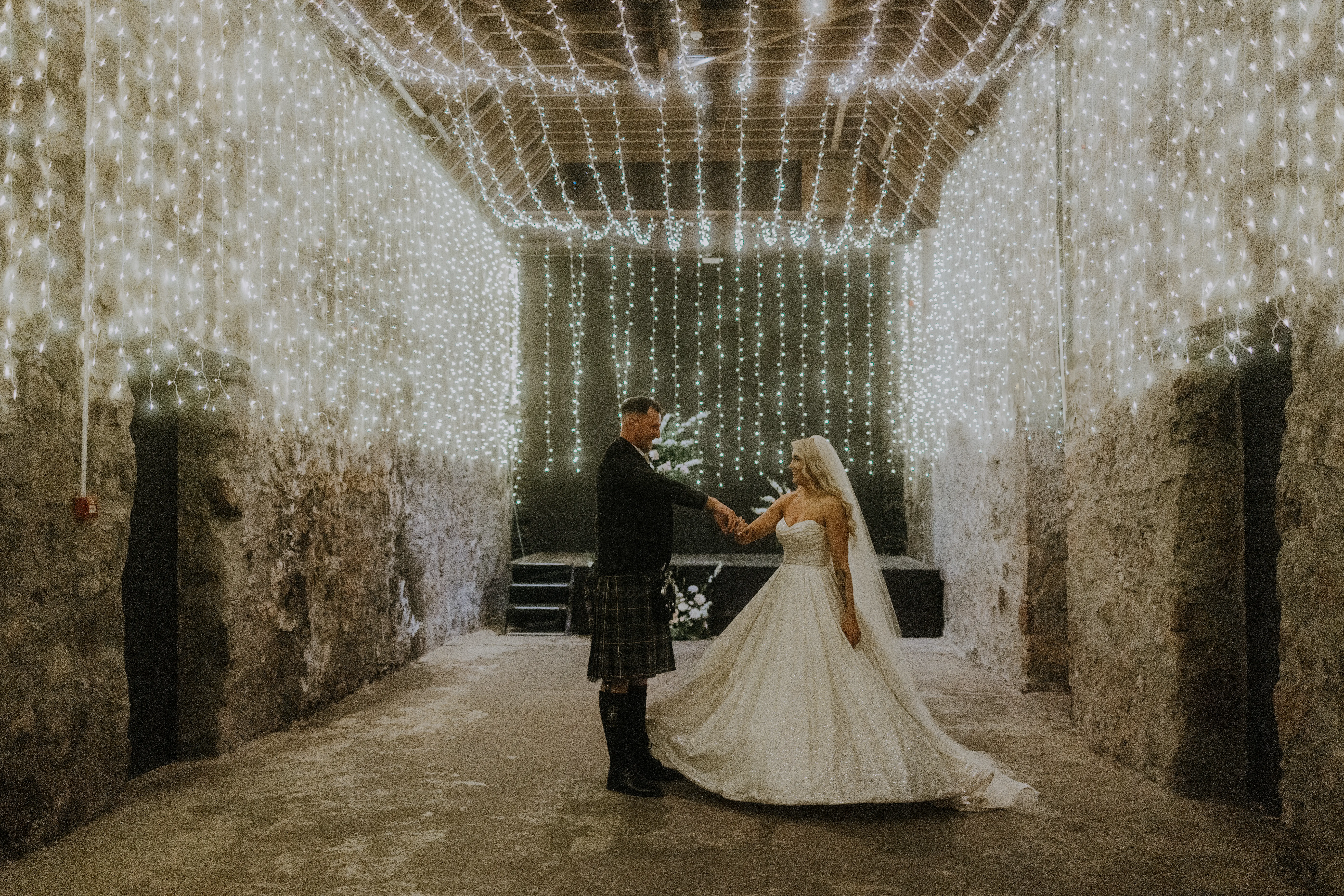 Bride Wearing Sparkly Aline Wedding Dress Called Anniston With Groom And Lights Hanging Throughout The Room