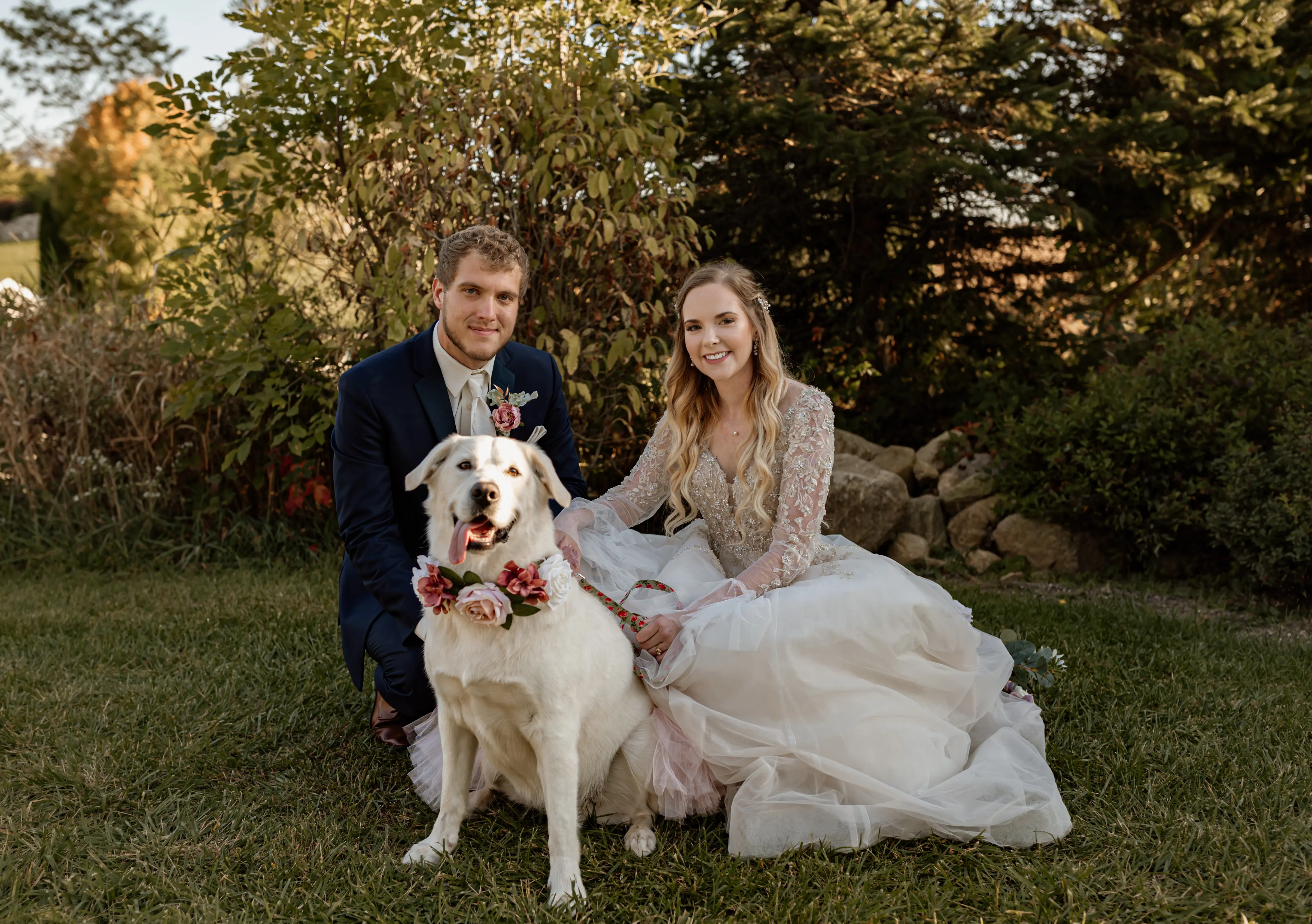 Bride, groom and dog sat in the grass on wedding day.