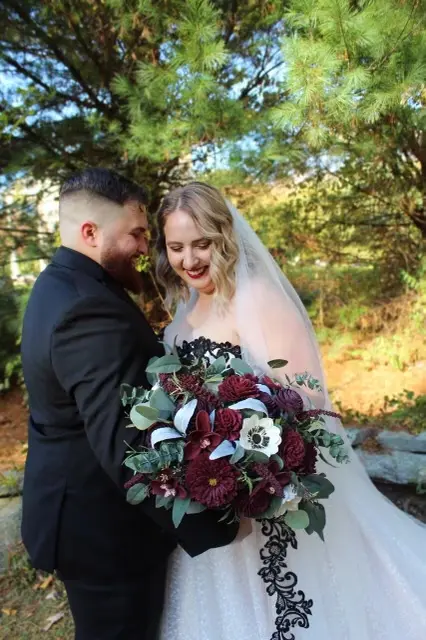 Bride Wearing Black Lace Wedding Dress Called Tristyn By Maggie Sottero With Groom Holding Maroon And White Flower Veil