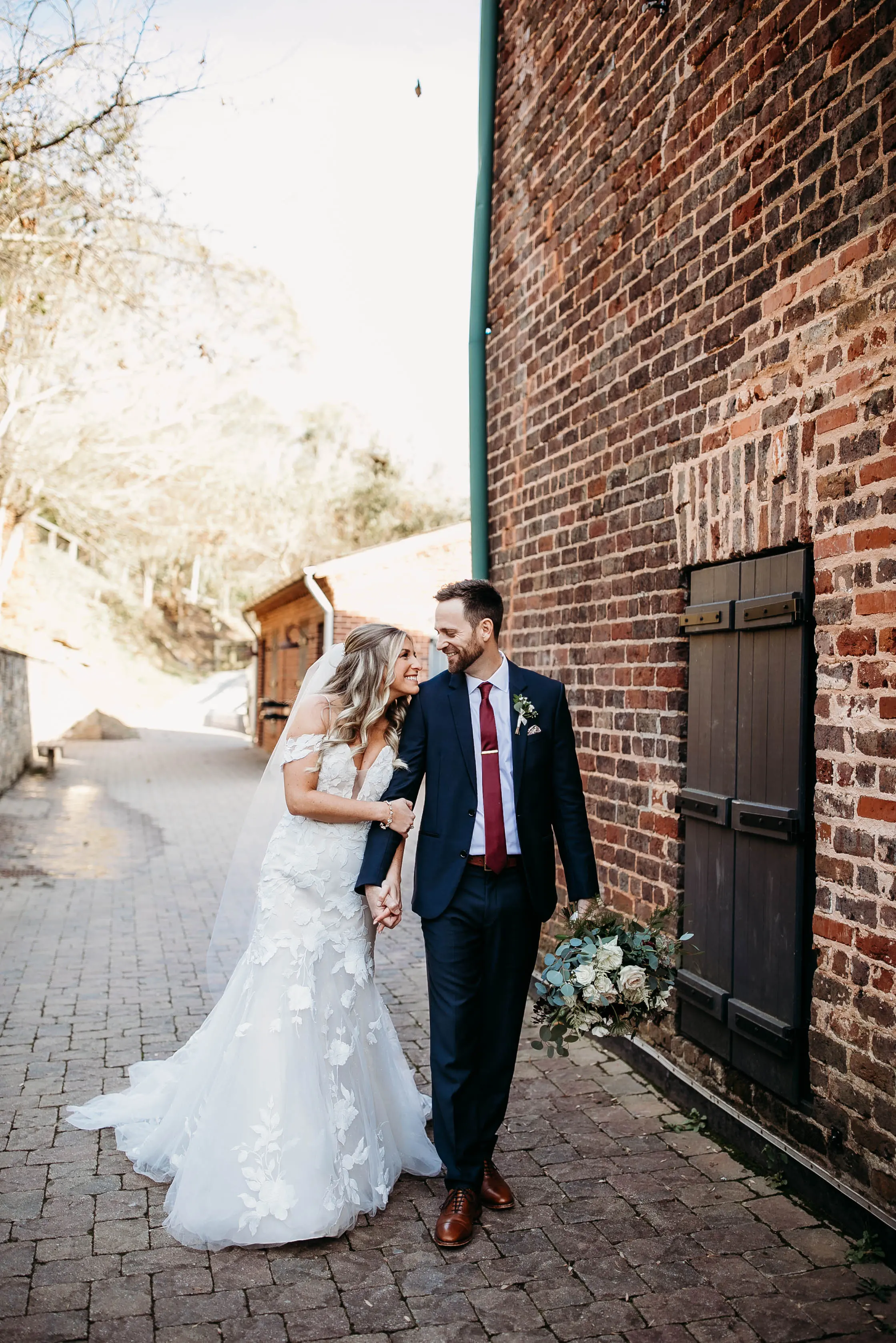Bride And Groom Walking Together. Bride Wearing Mermaid Wedding Dress With Oversized Lace Called Hattie By Rebecca Ingram