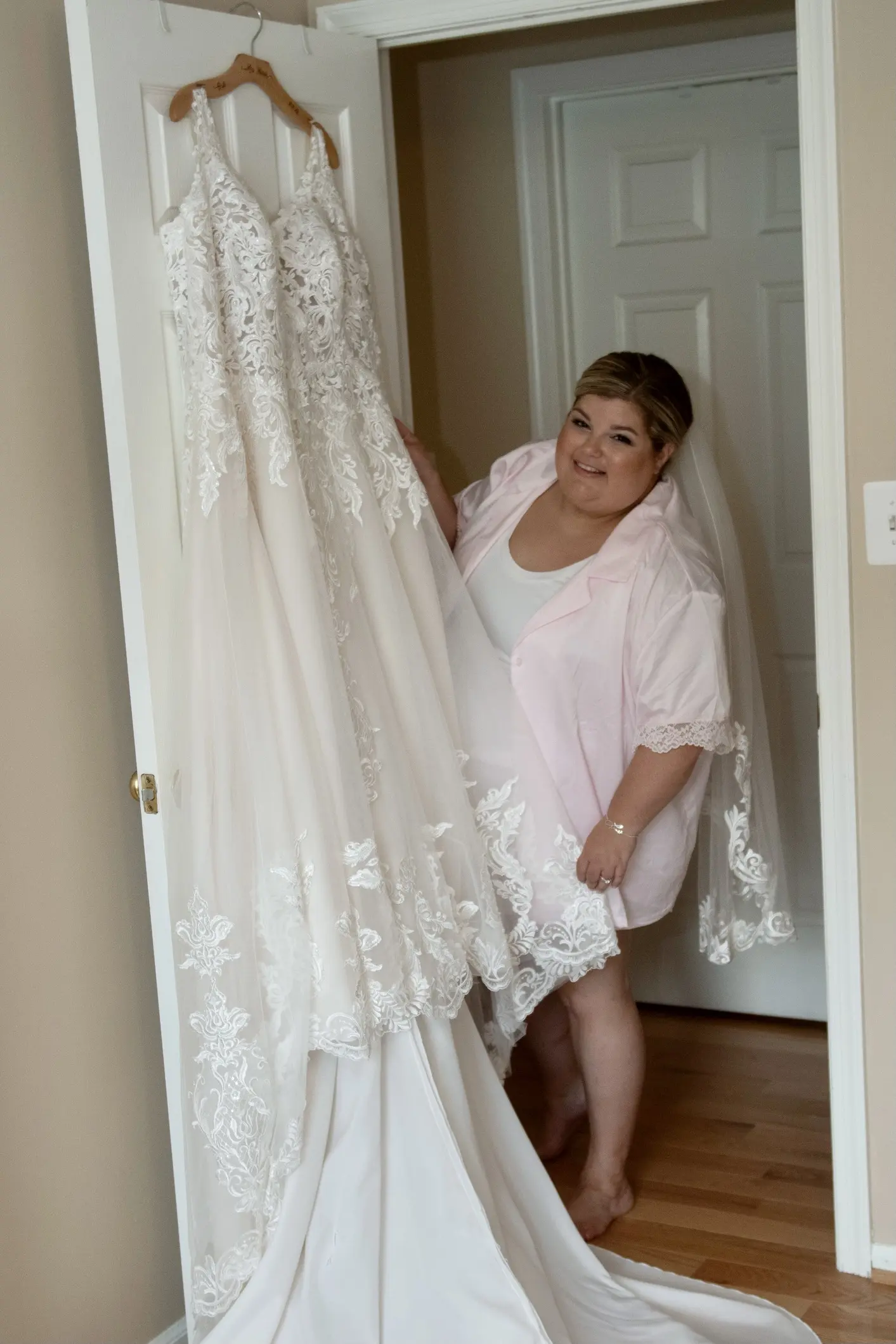 Bride standing next to her hanging Lace A-line Wedding Dress called Trinity Lynette by Maggie Sottero