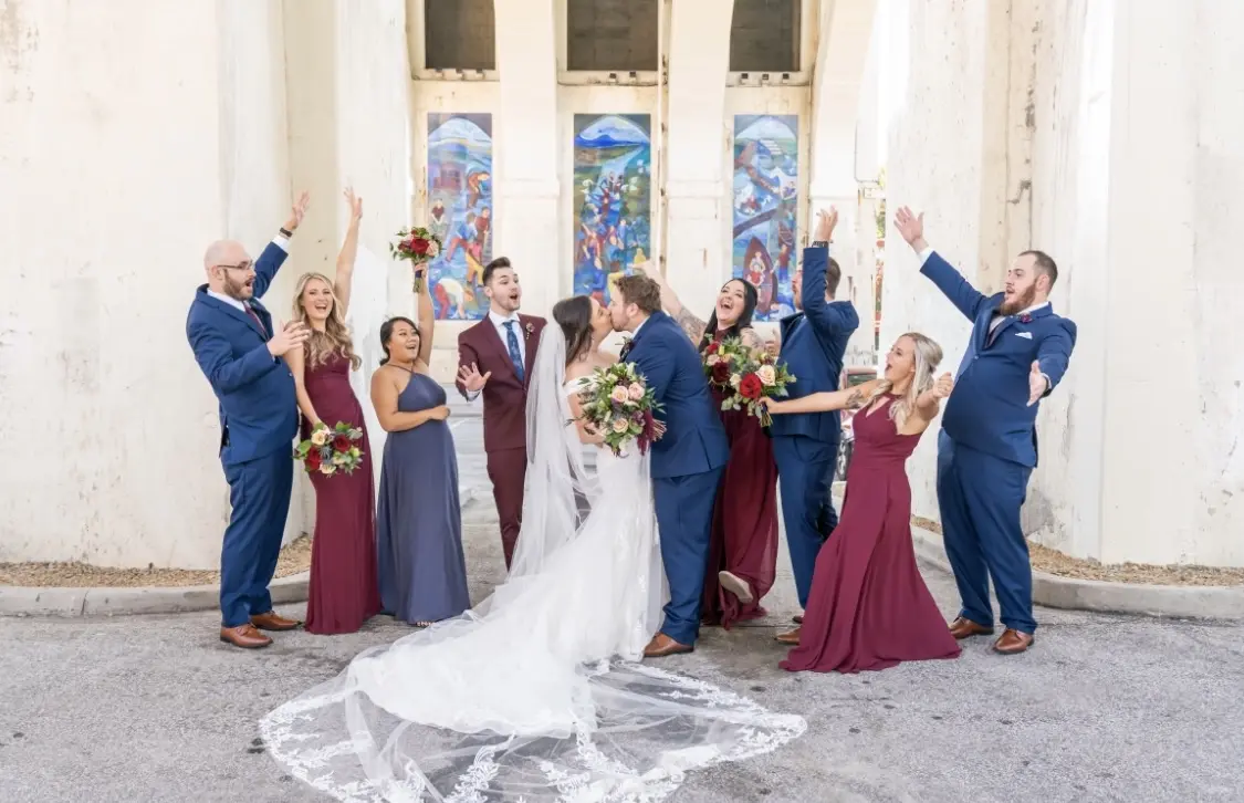 Bride wearing cap sleeve lace mermaid wedding dress while kissing groom in front of bridal party.