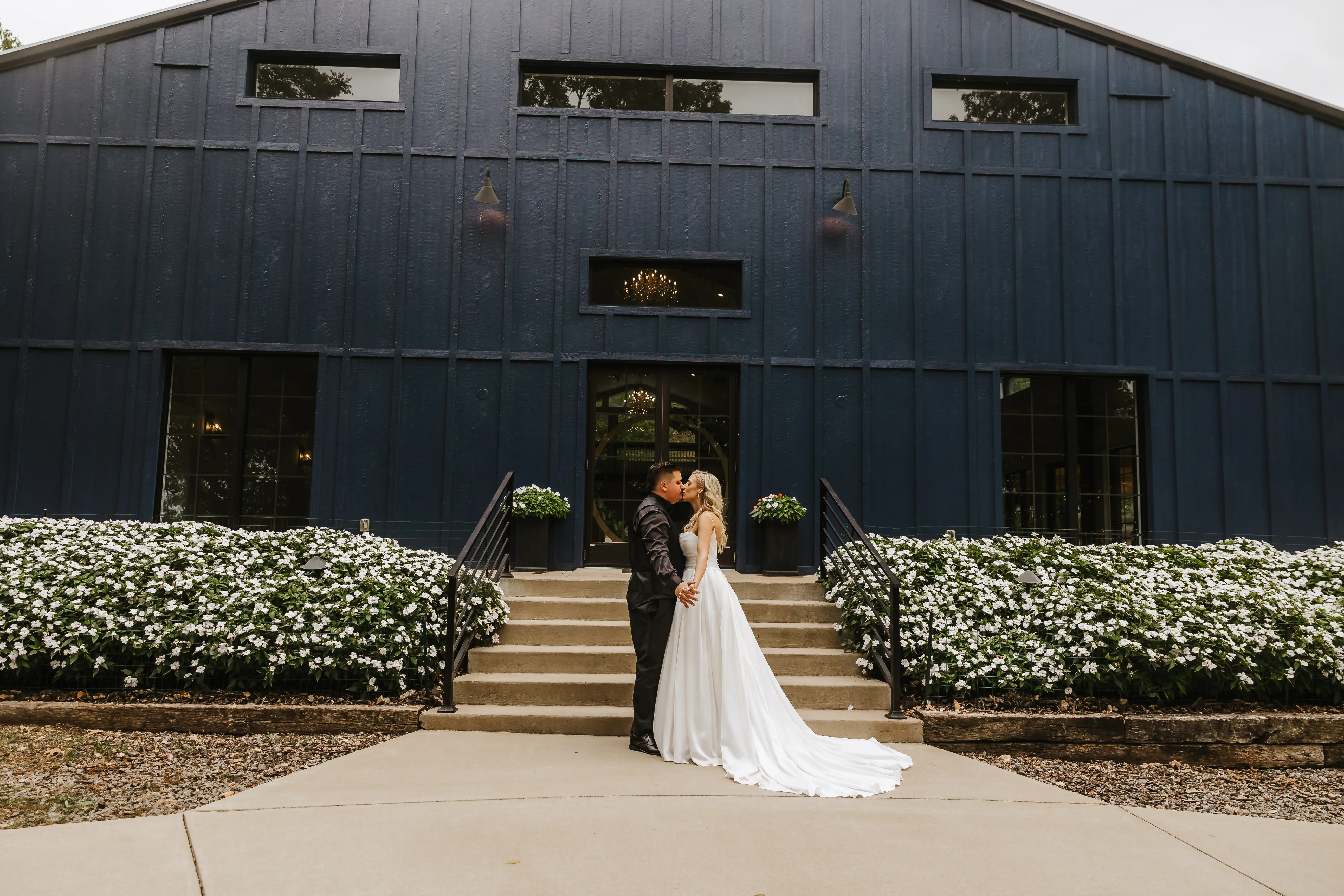 Bride Wearing Aline Satin Wedding Gown Called Scarlet By Maggie Sottero Kissing Groom In Front Of A Black Building
