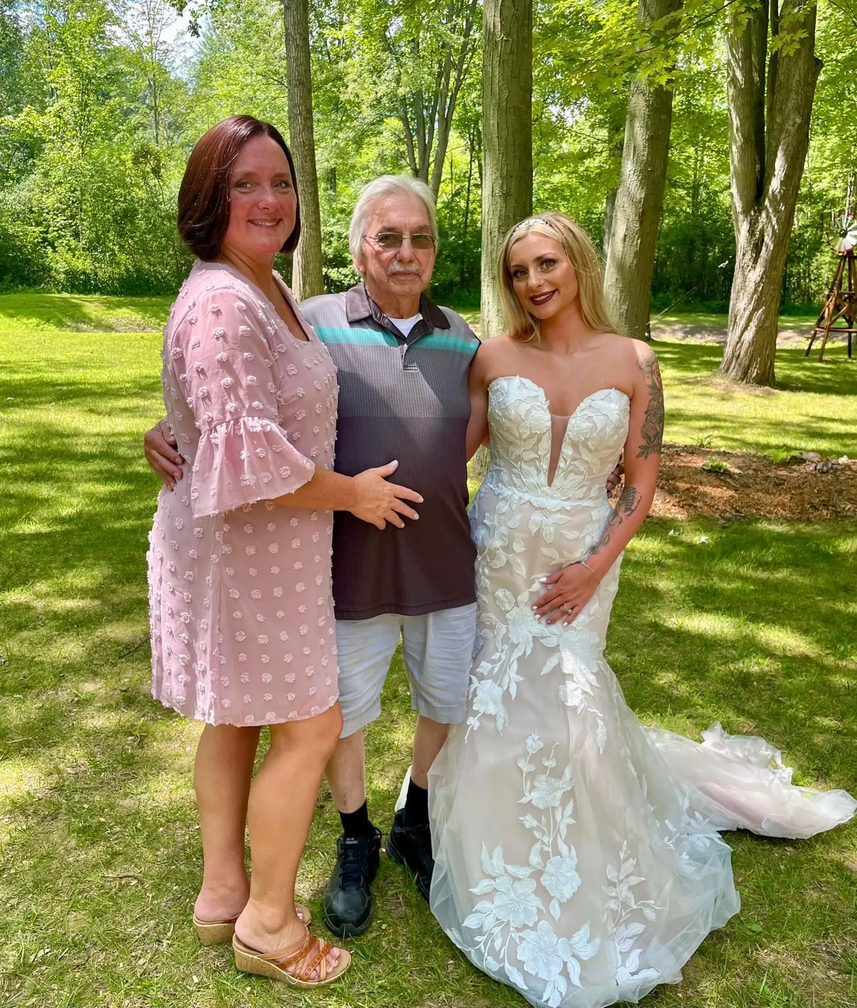 Bride Wearing Lace Strapless Mermaid Wedding Dress Called Hattie By Rebecca Ingram Posing With Parents On Green Grass