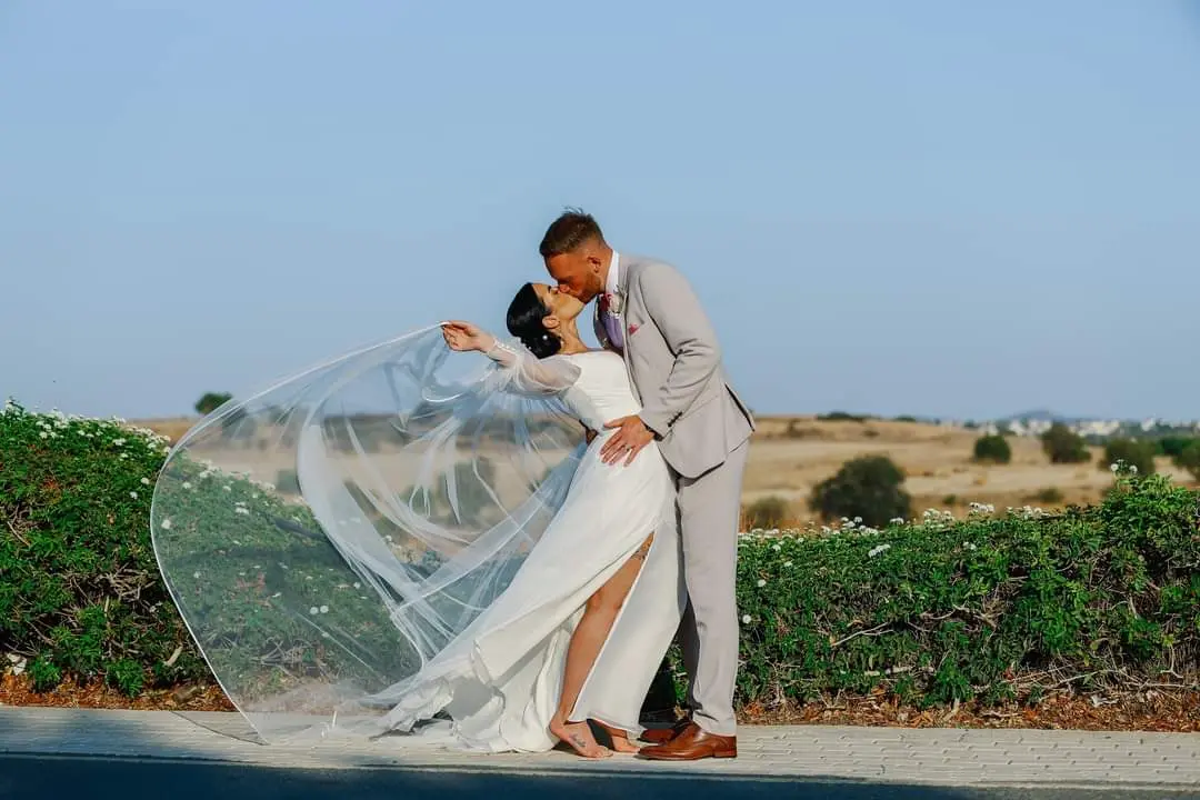 Bride Wearing Boho A-Line Wedding Dress Called Nerida By Sottero And Midgley Throwing Veil In The Wind While Kissing Groom
