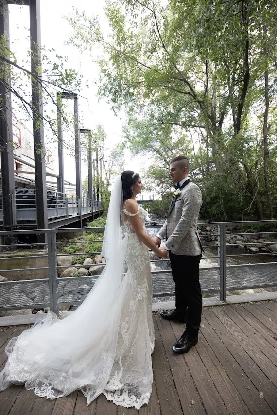 Bride Wearing Lace Sheath Wedding Dress Called Tuscany Lynette By Maggie Sottero With Groom On A Bridge Over A River
