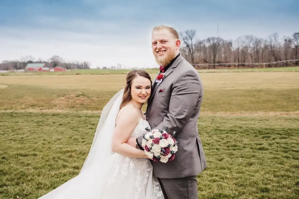 Bride And Groom Standing Together In A Field. Bride Wearing Lace Tulle Wedding Dress Called Stevie By Maggie Sottero