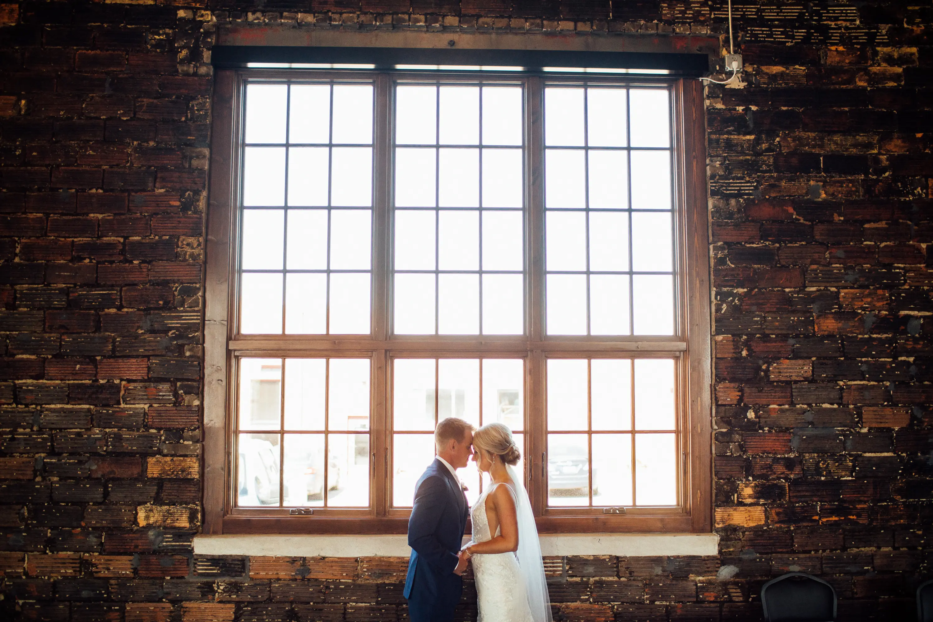 Bride Wearing Graceful Sparkly Lace Wedding Dress Called Fontaine By Maggie Sottero With Groom In Front Of Large Window