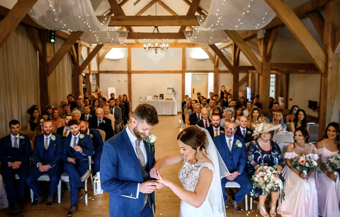 Bride placing ring on her husband to be with wedding guests in the background.